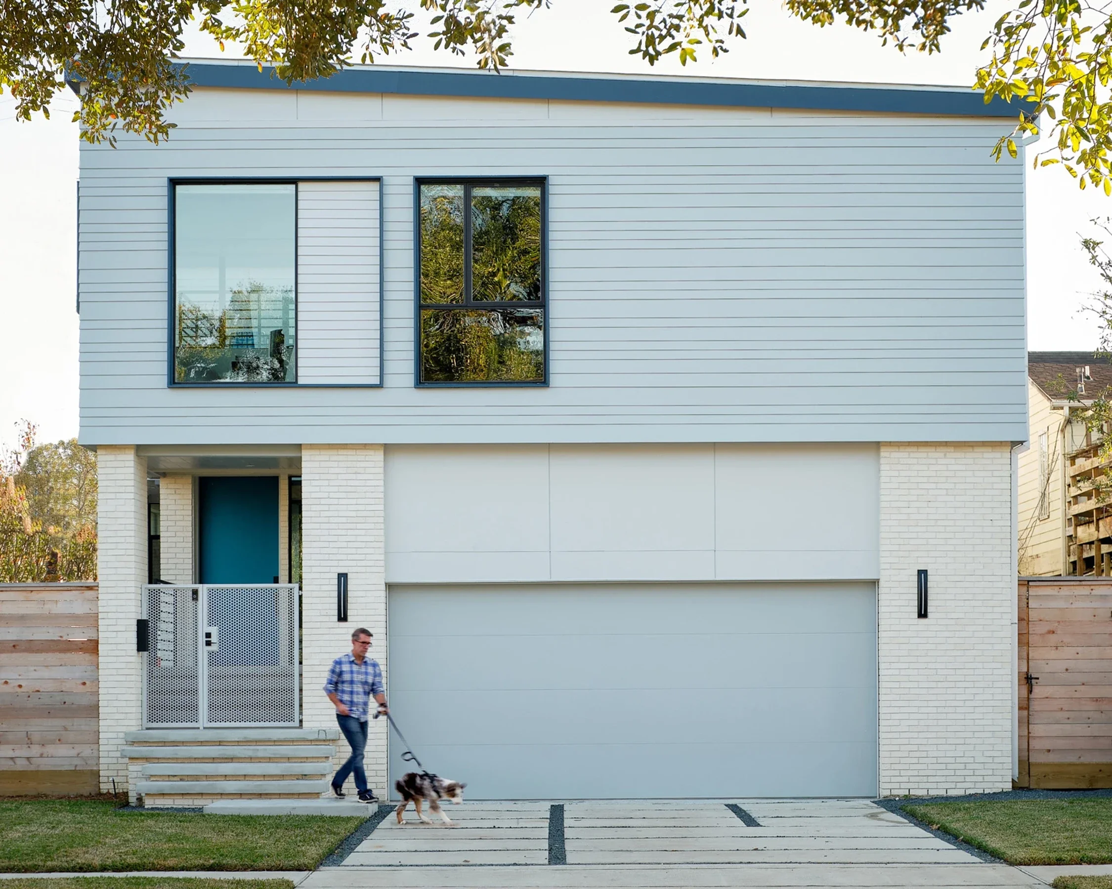 A modern two-story house with white brick and siding exterior, large windows, and a white garage door. A man in a blue plaid shirt walks on the driveway with his dog.