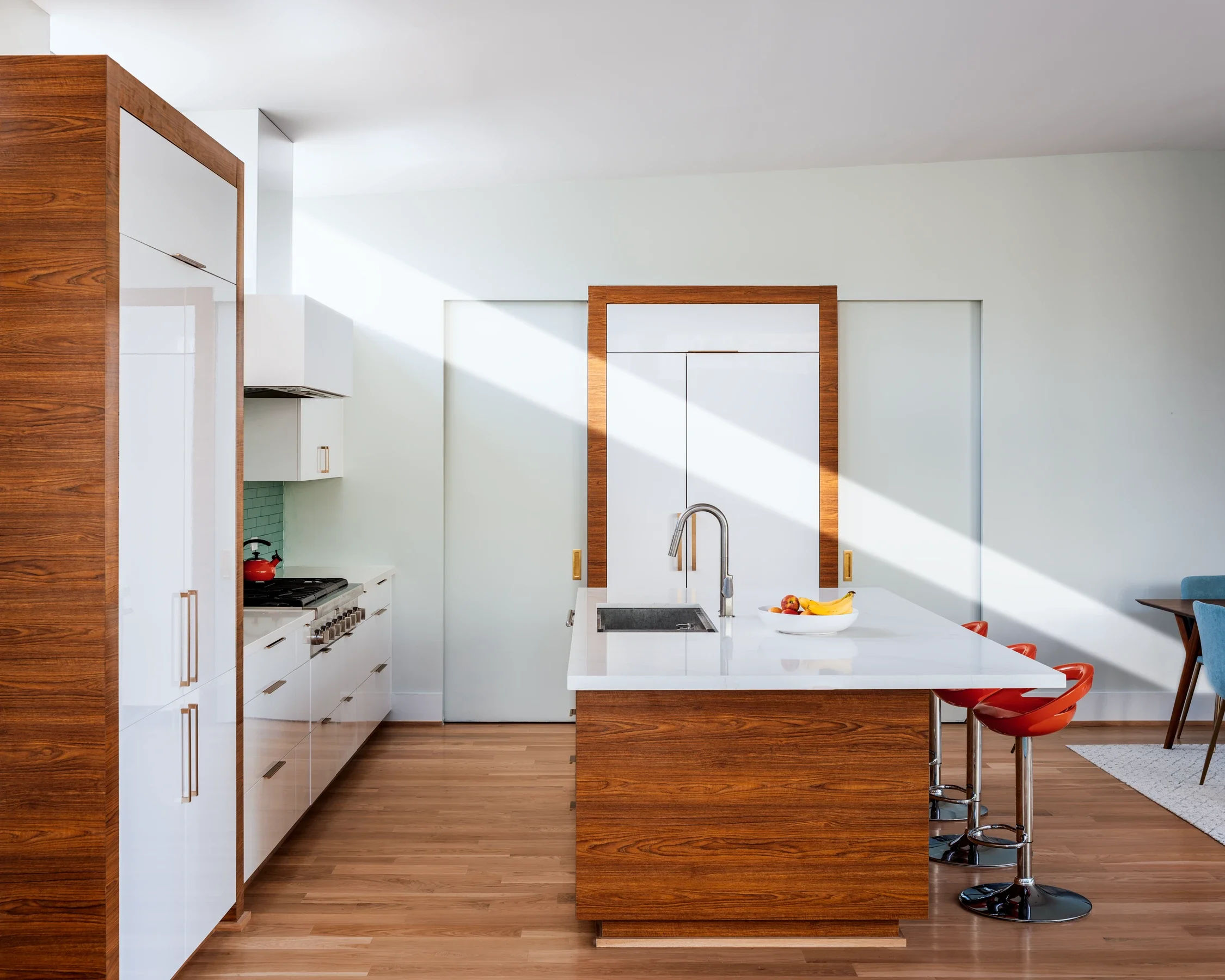 Modern kitchen with white cabinets, wooden accents, an island with a white countertop, a sink with a faucet, and a bowl of fruit, illuminated by sunlight.