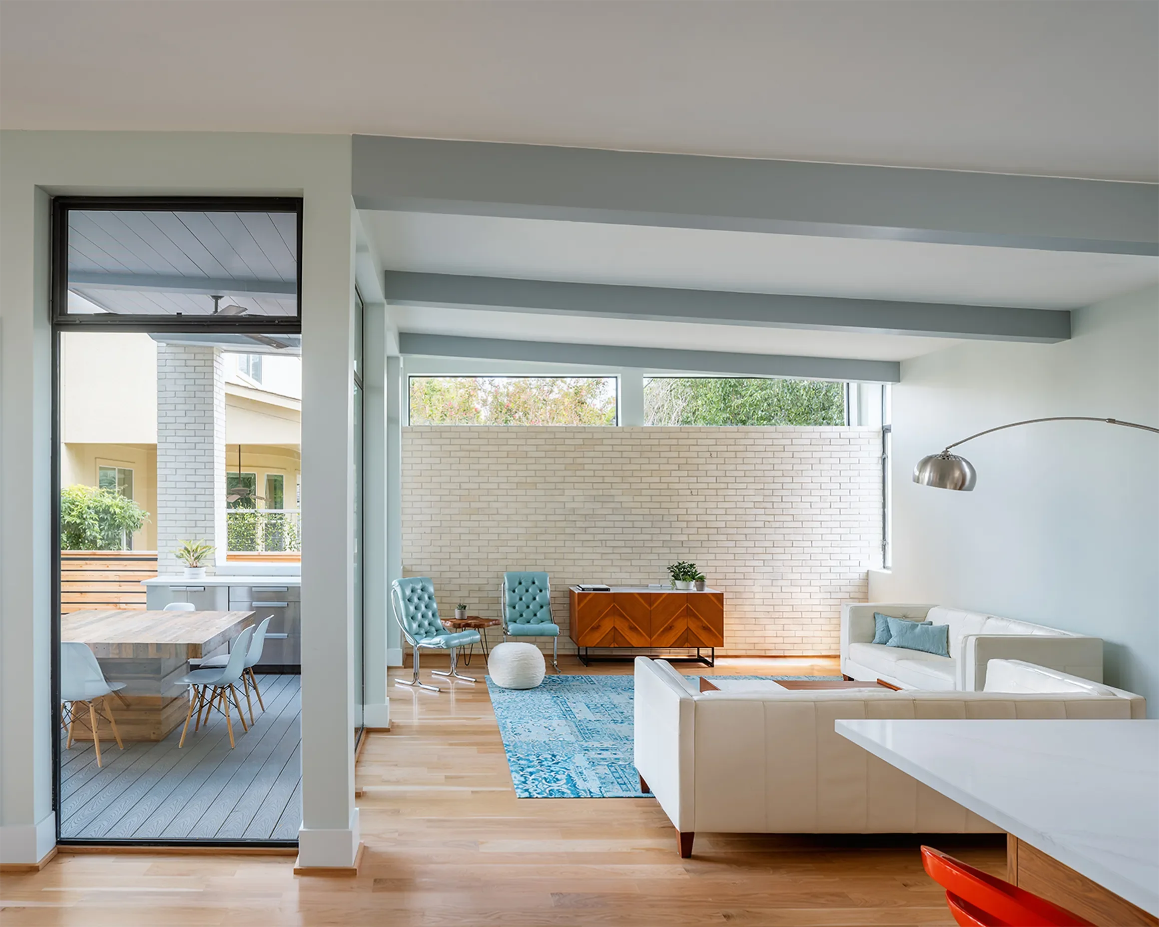 Modern living room with white sofa, two turquoise armchairs, a wooden sideboard, a blue rug, and a white brick wall, with large windows and an adjacent dining area.