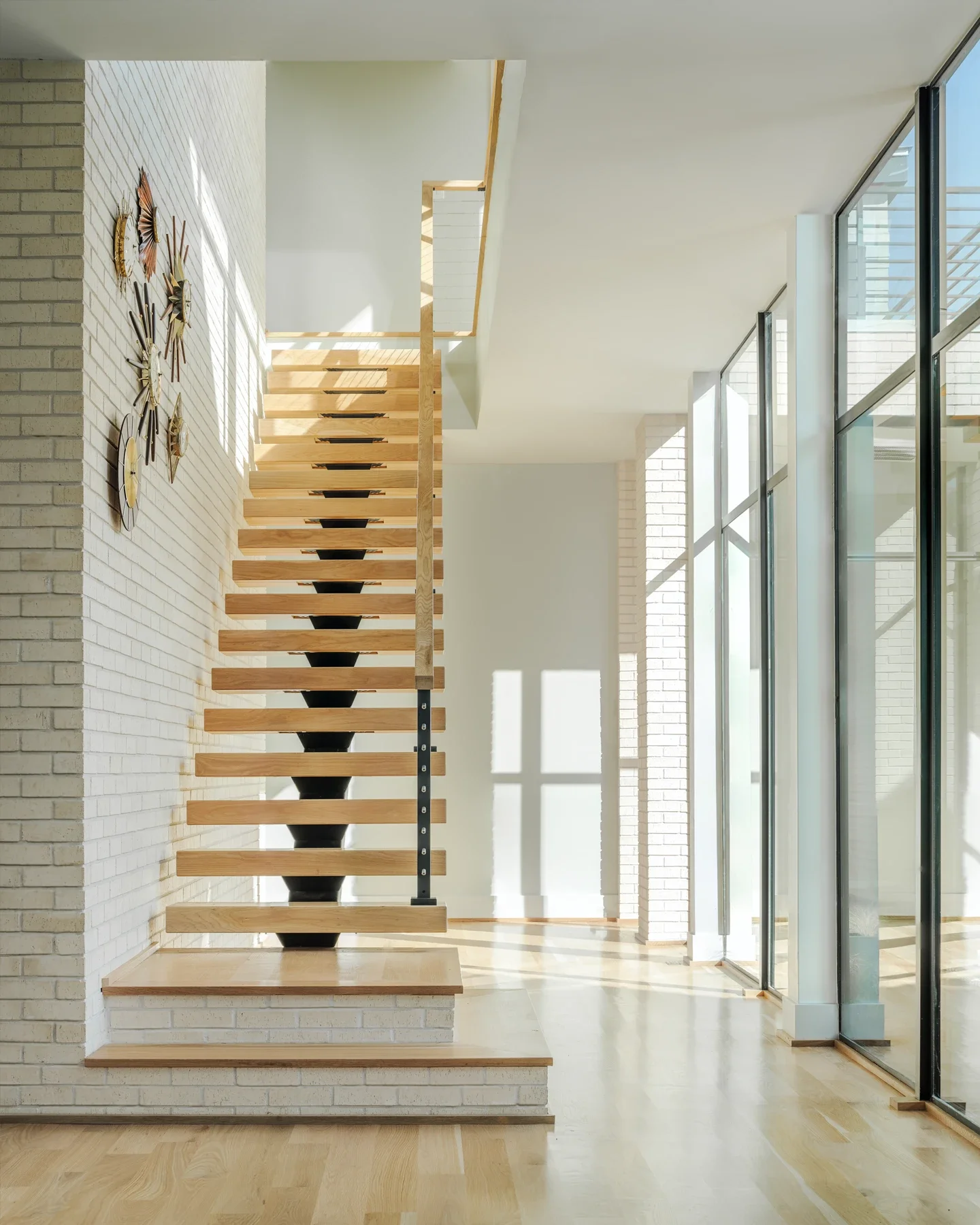 Interior of a modern house with a floating wooden staircase, white brick walls, large floor-to-ceiling windows, and natural light.