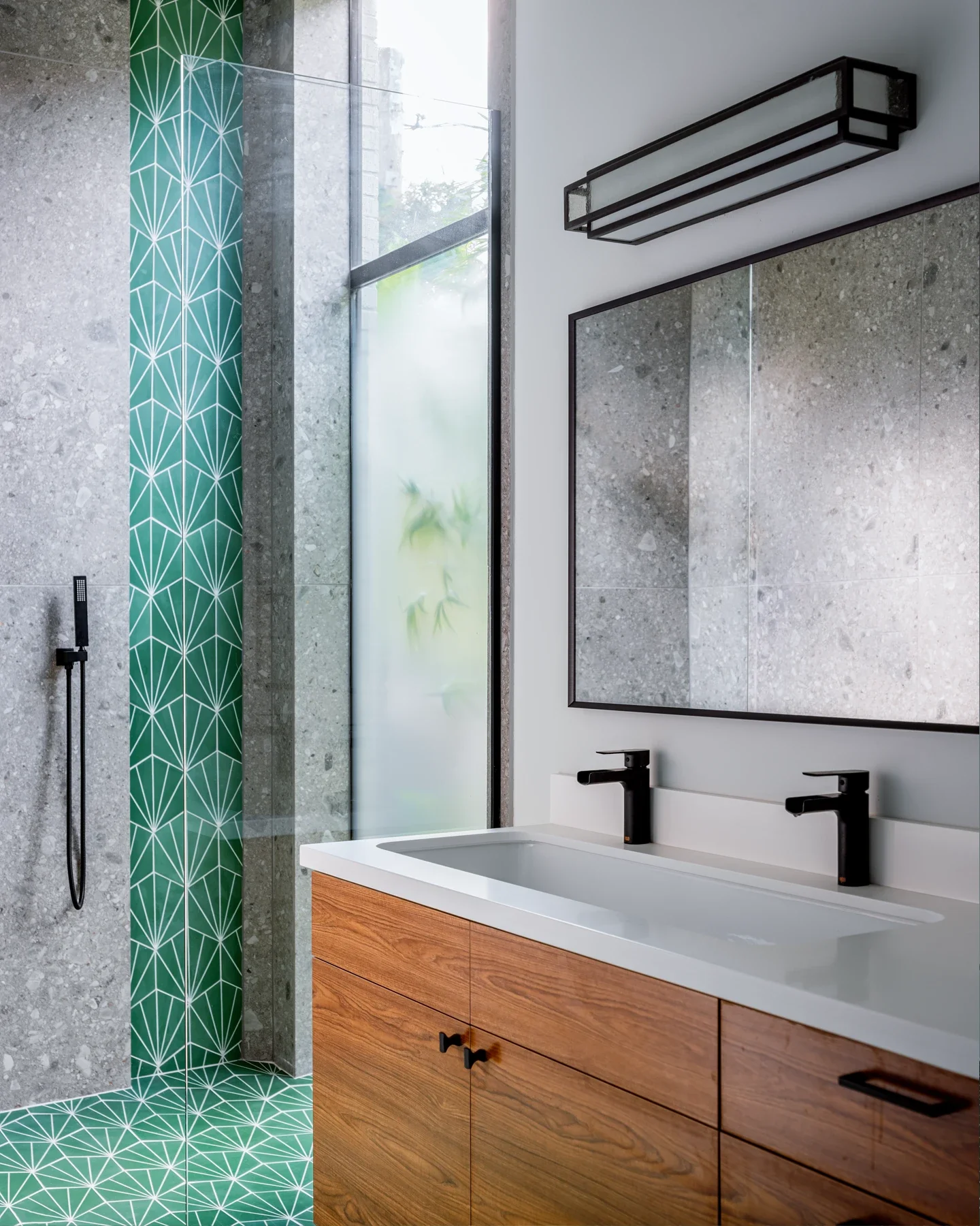 Modern bathroom with a large mirror, dual black sinks, wooden cabinet, and a shower area with textured concrete and green geometric tiled wall and floor.