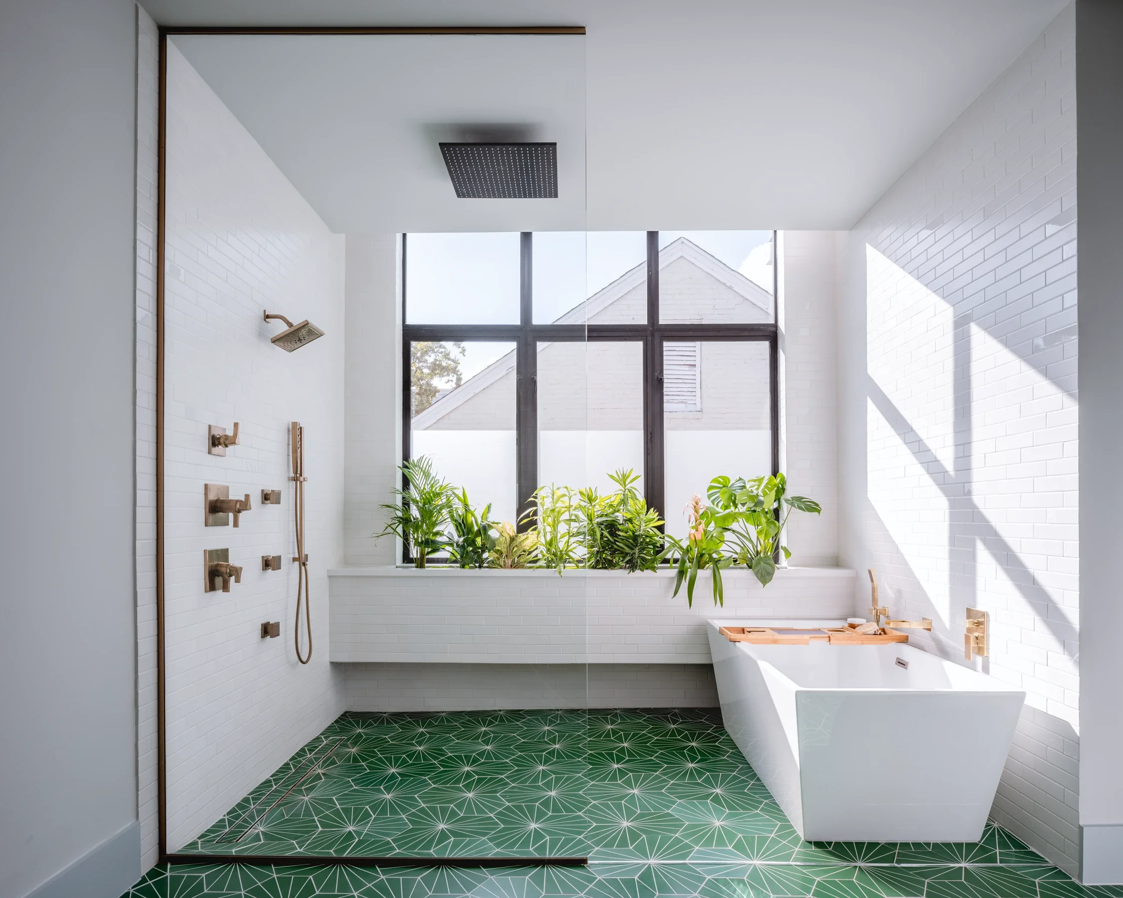 Modern bathroom with white brick walls, green geometric tile floor, large window with plants, walk-in shower with brass fixtures, and white bathtub.