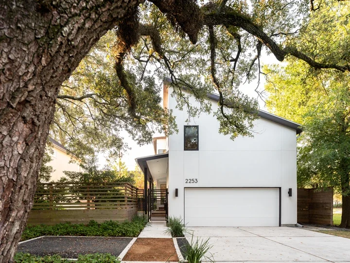 Front view of a modern two-story house with a white exterior, black accents, a garage door, and a pathway leading to the entrance, surrounded by trees and greenery.