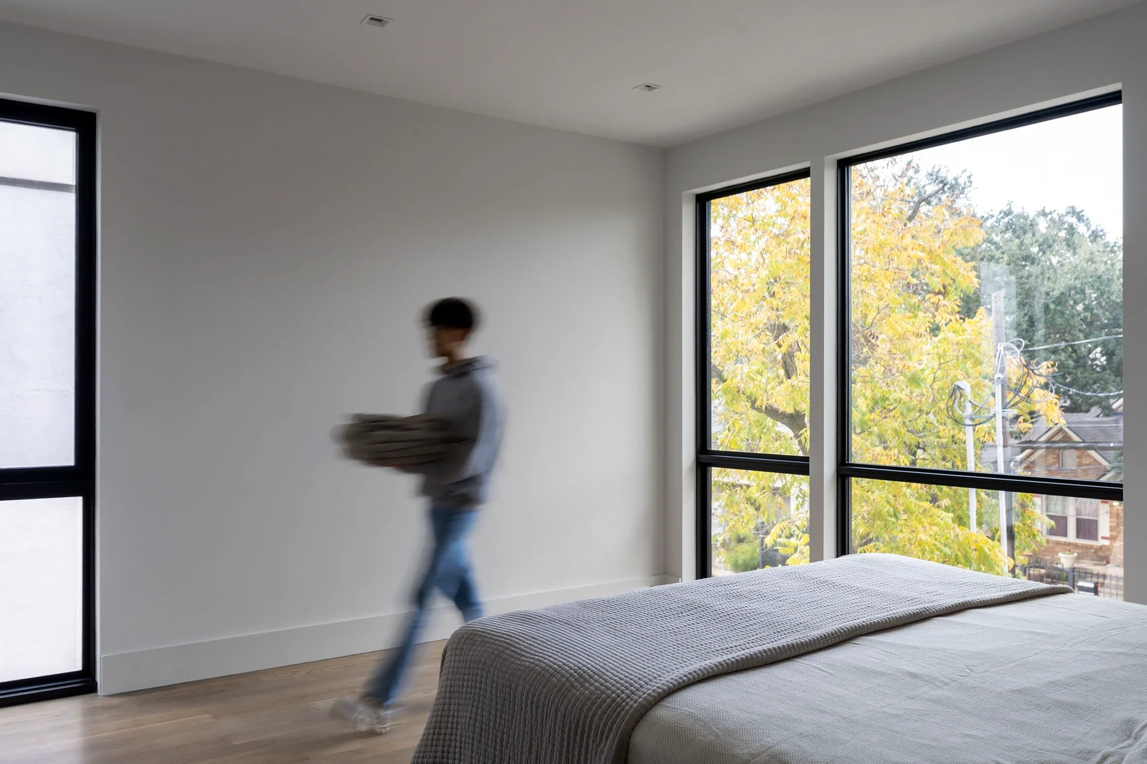 Person walking across a bedroom with large windows showing autumn trees outside.