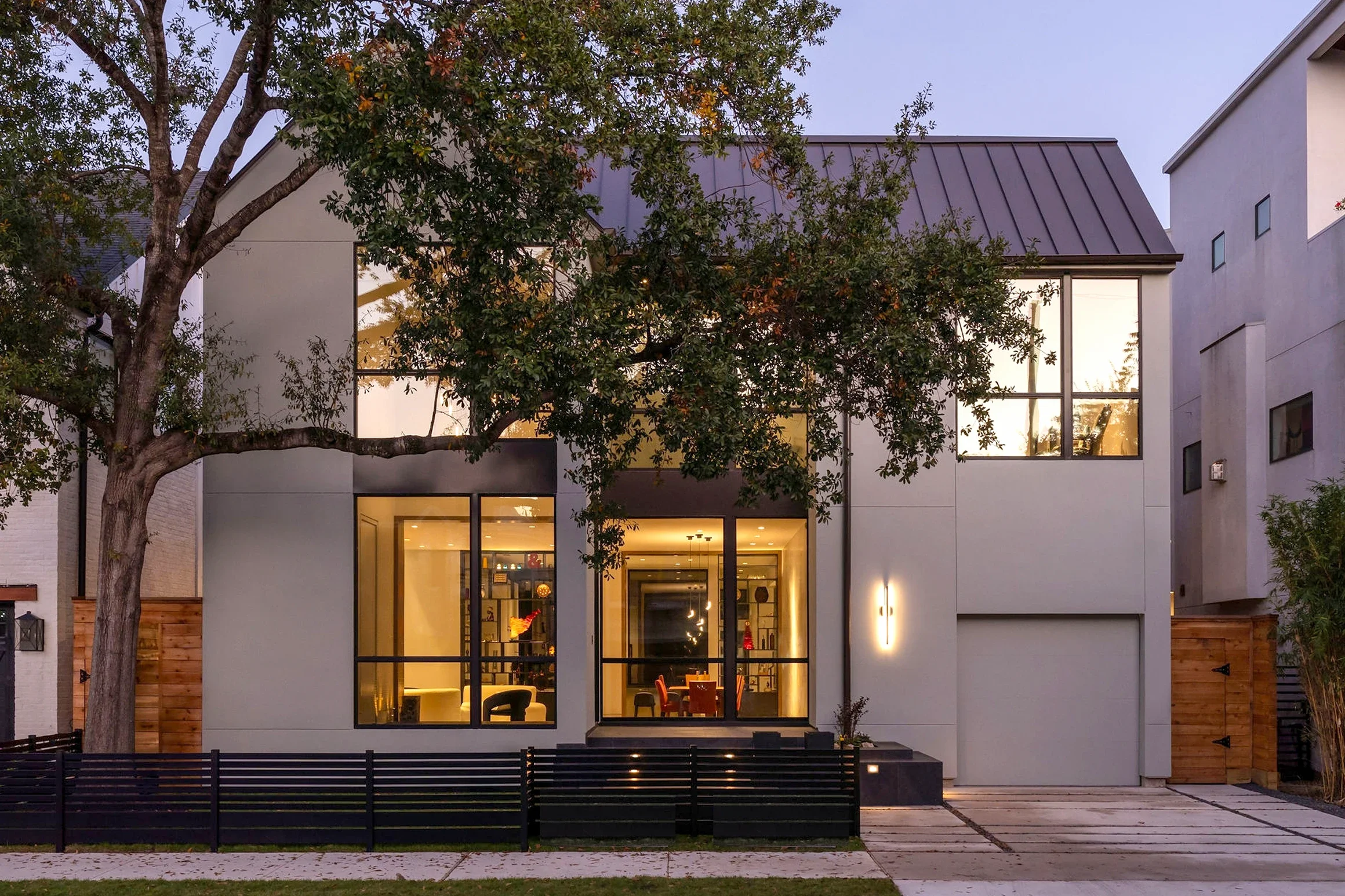 Modern two-story house with large windows, black metal roof, and front driveway, surrounded by trees and fencing in the evening.