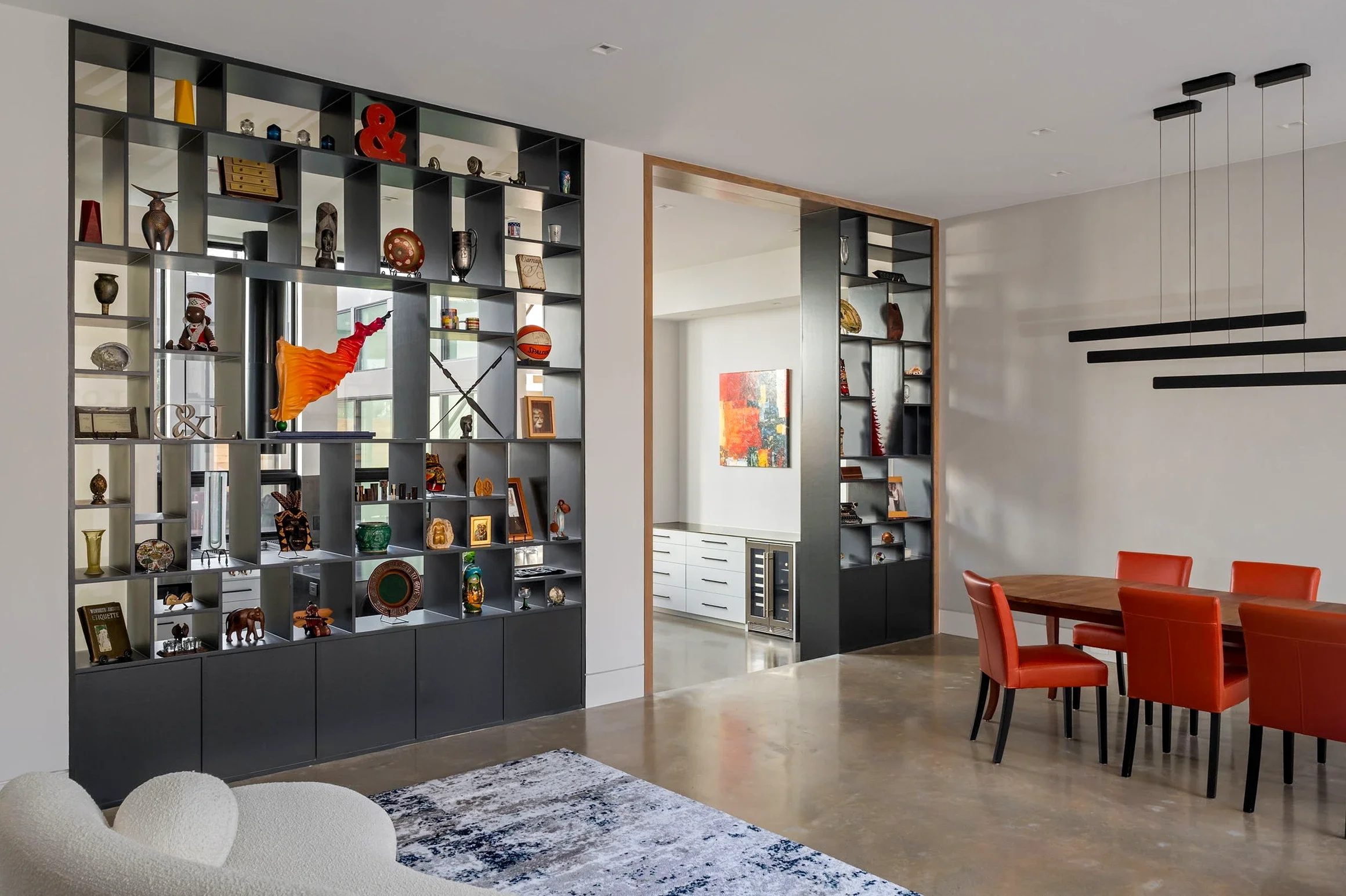 Open concept living and dining room with tall black numeric display shelf filled with decorative objects and art, adjacent to a white wall with three black horizontal light fixtures, a wooden dining table with six orange chairs, and a partial view of