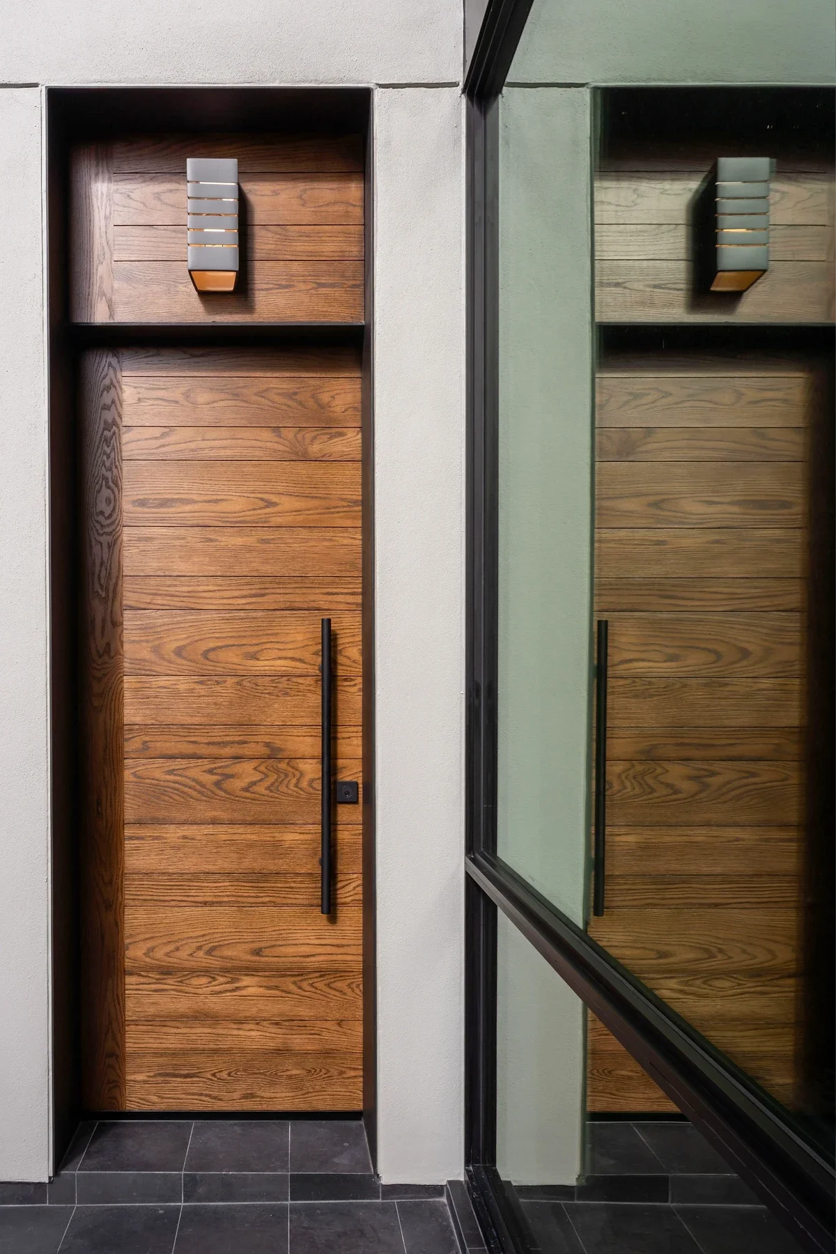 A wooden door with a long black handle, set in a modern building facade. The door has a small rectangular light fixture above it. To the right, there's a glass window reflecting the door and part of the building.