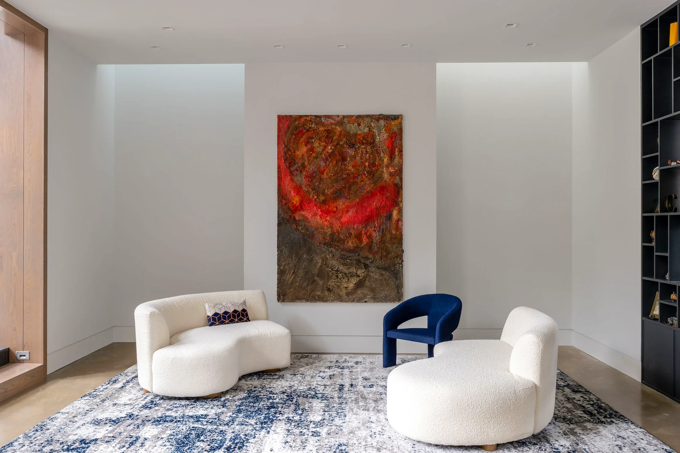Modern living room with a white curved sofa, a blue armchair, a beige armchair, a large abstract red and brown painting on the wall, a black shelving unit on the right, a patterned rug on the floor, and white walls.