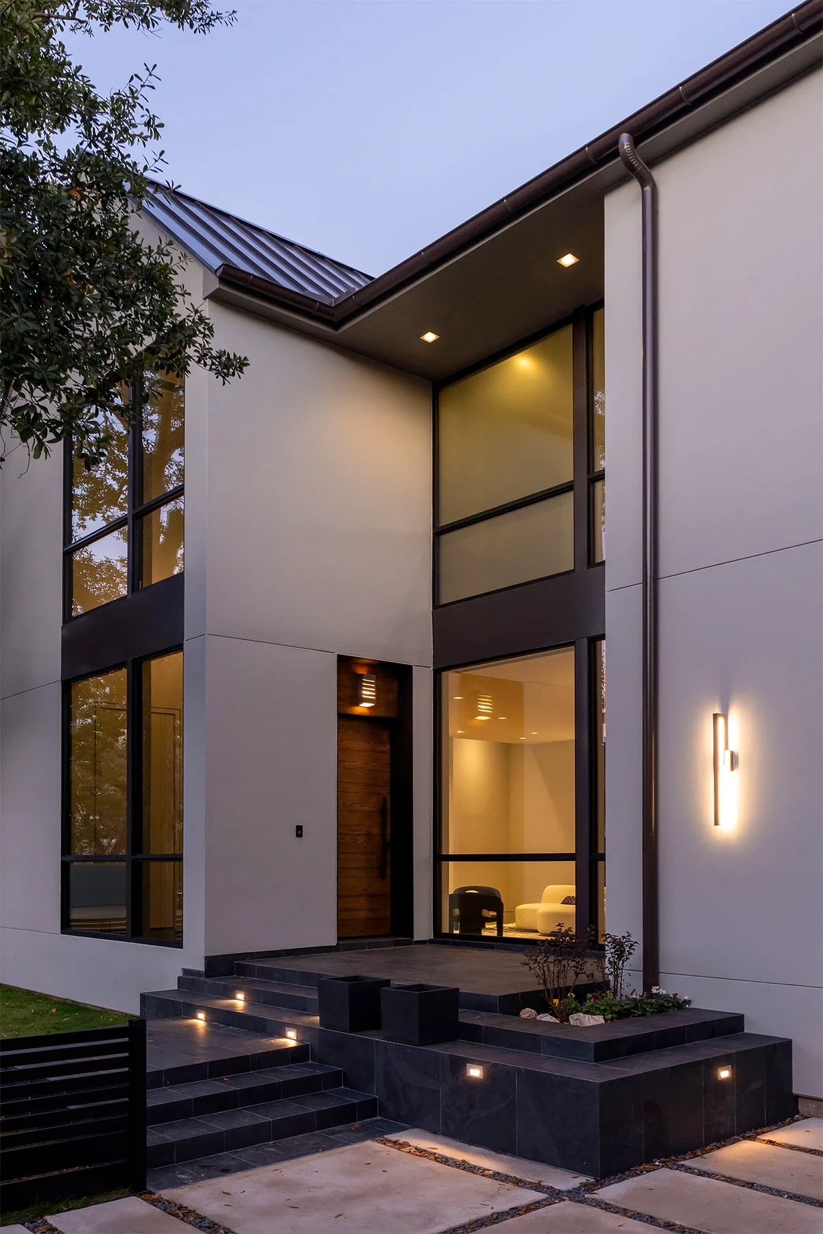 Modern house with large windows, exterior lighting, a wooden front door, and a small landscaped porch area with steps and pathway