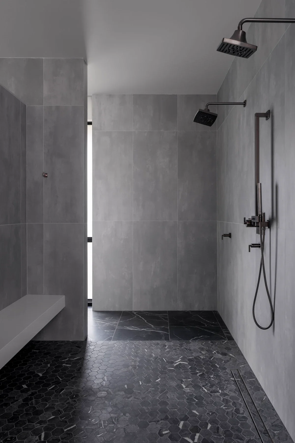 A modern, minimalistic walk-in shower with grey tile walls and black honeycomb floor tiles, featuring two overhead rainfall showerheads and a handheld shower wand.