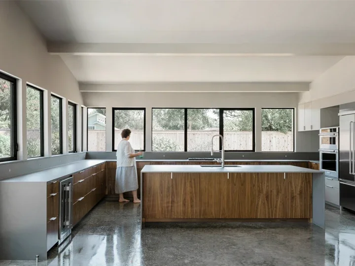 A woman standing in a modern kitchen with large windows, wooden cabinetry, and a central island, looking out the window.