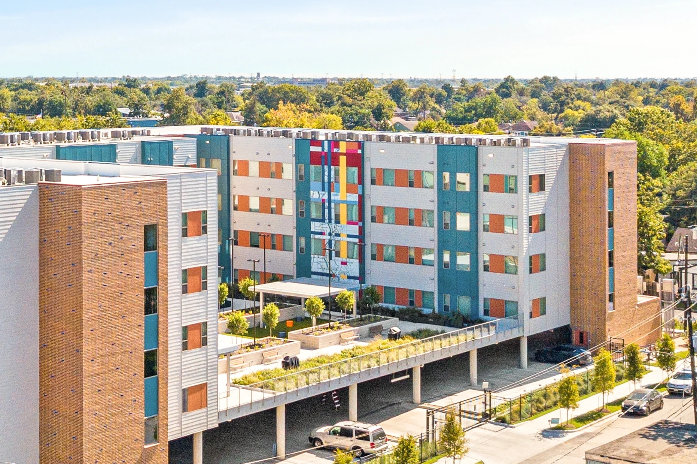 Modern residential apartment complex with colorful mural, trees, and parked cars in the foreground.
