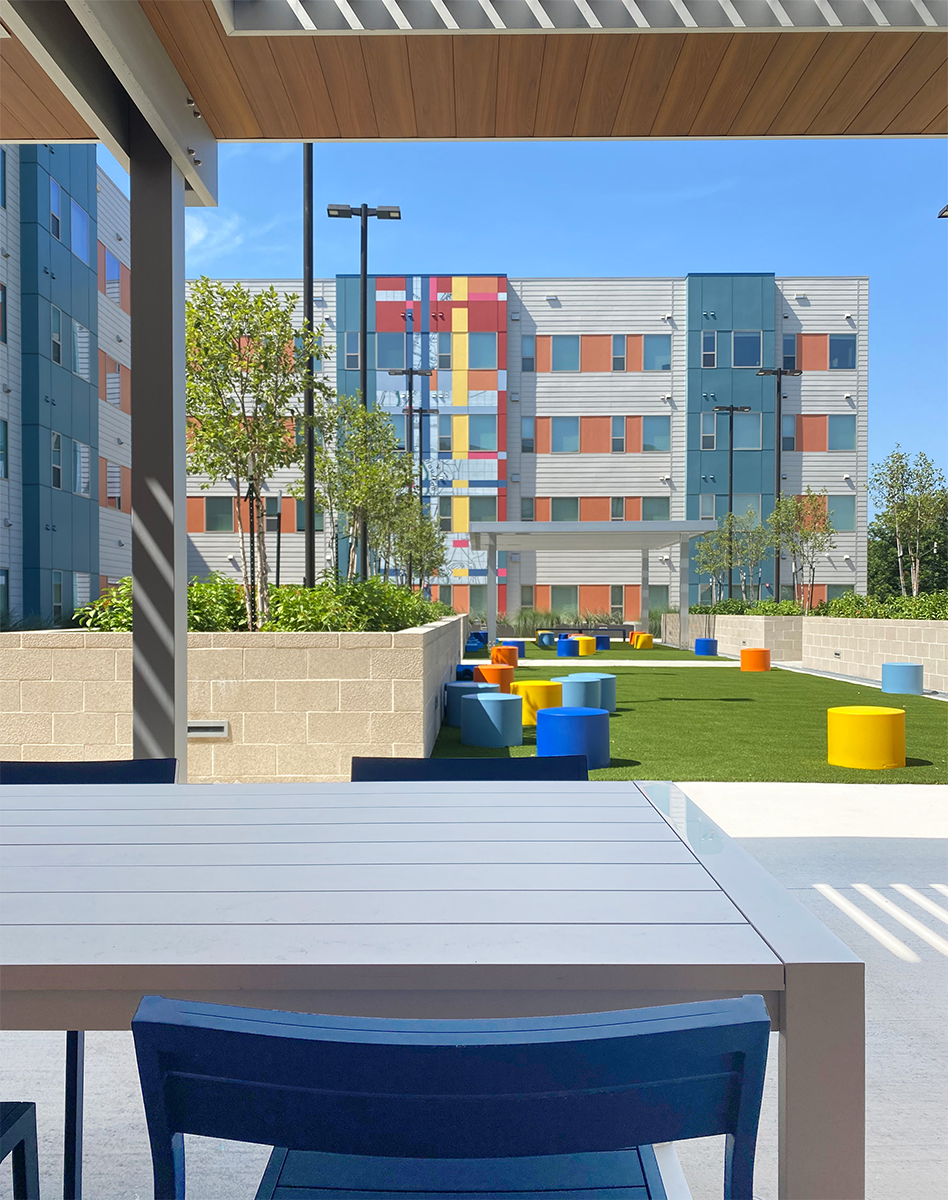 Outdoor seating area with a white table and blue chairs under a roof, overlooking a grassy courtyard with colorful cylindrical seats, trees, and a modern multi-story building with a colorful facade.