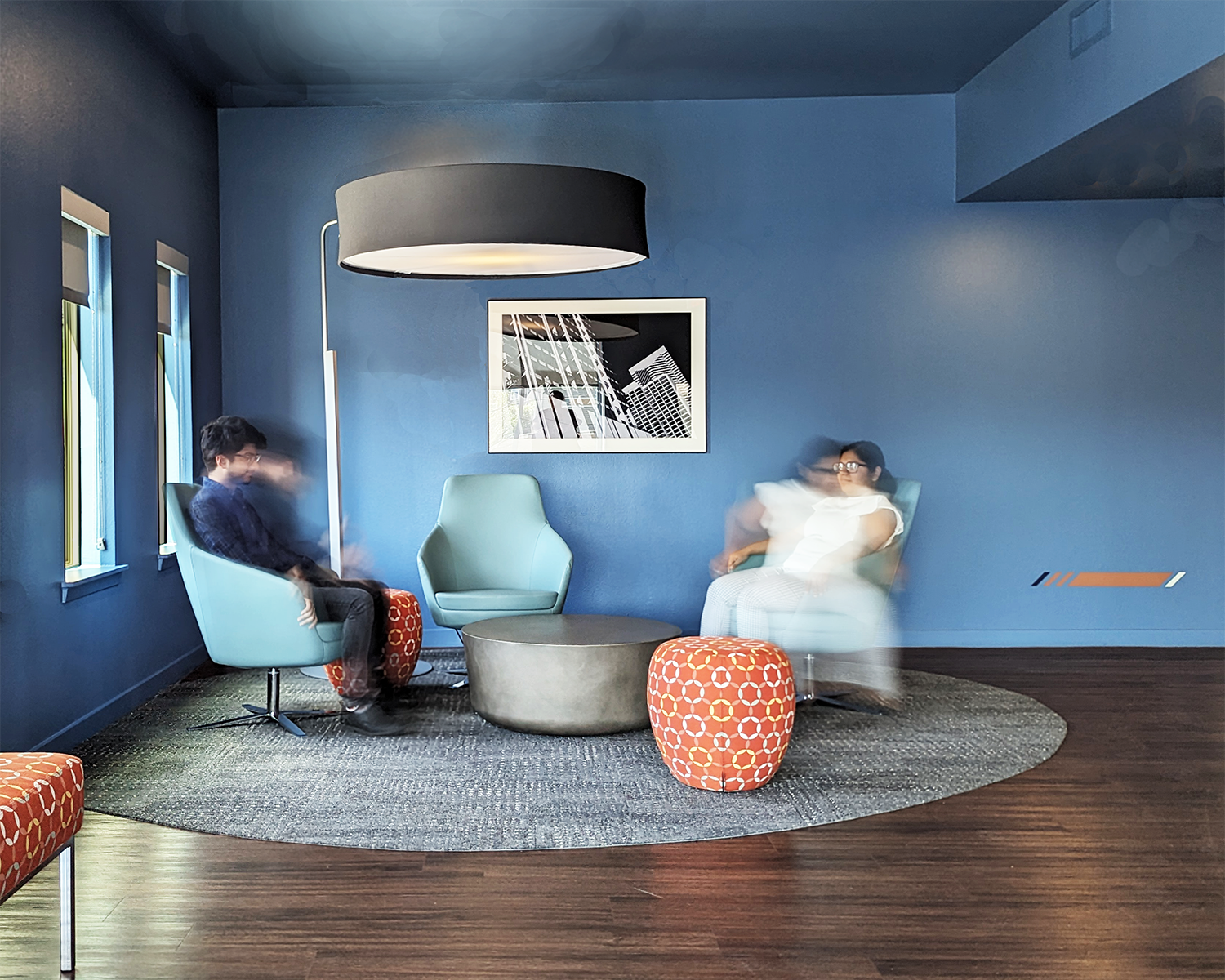 Modern living room with blue walls, two women and one man sitting in chairs with blurred motion, a round coffee table, patterned ottomans, framed black and white architectural photograph, large black pendant light.