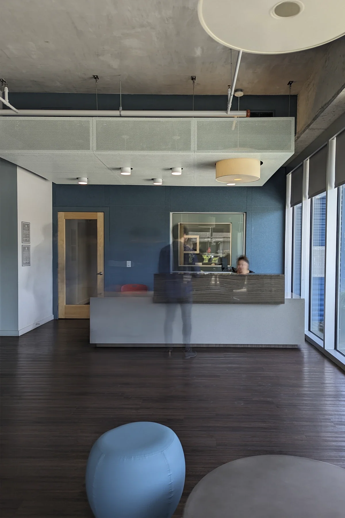 Modern office reception area with a reception desk, blue wall behind it, and large glass windows to the right. There are two people at the desk, with one standing and one seated, and some furniture in the foreground, including a blue cushion and a ro