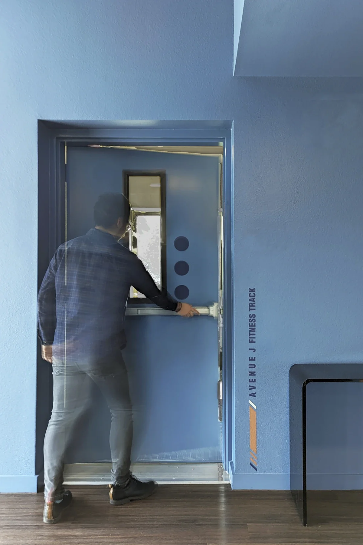 A man in a checkered shirt and gray pants is operating a wheelchair lift on a blue door at the Avenue E J Fitness Track. The door and surroundings are painted blue, with a black table or counter to the right.