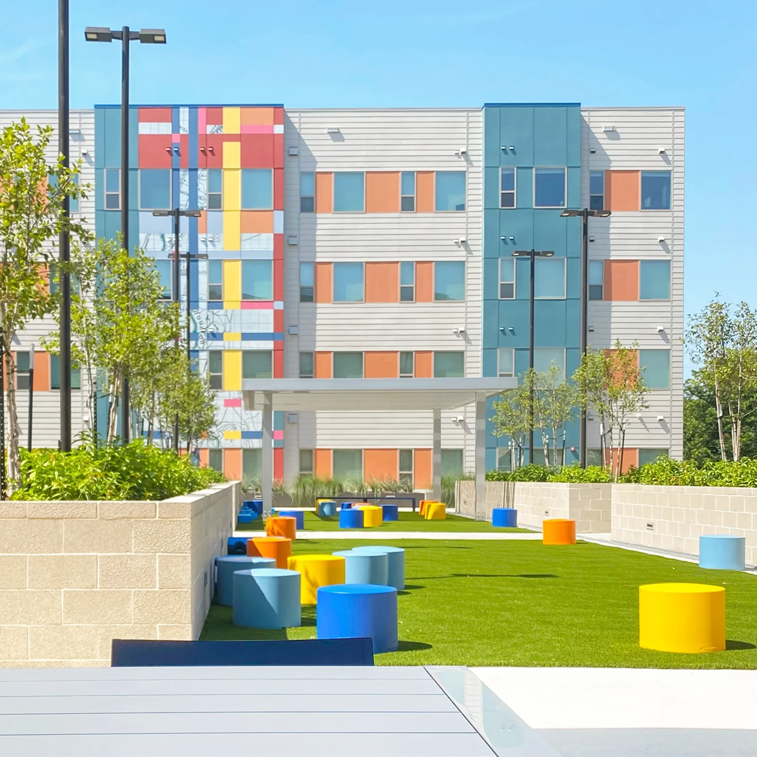 Outdoor space in front of a modern multi-story building with colorful accents, featuring a lawn area with scattered colorful round stools, trees, and a sheltered seating area.