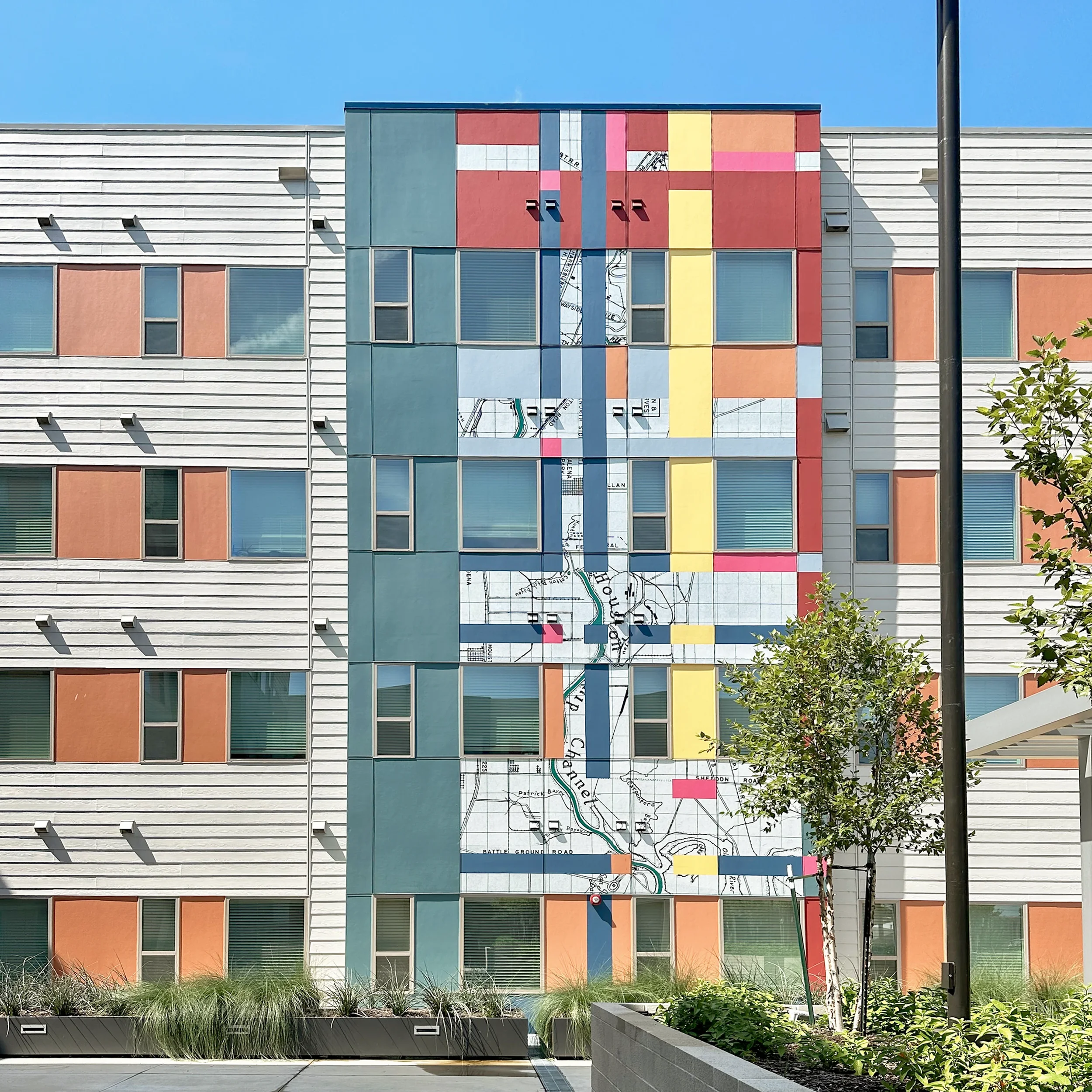 Colorful mural on the side of a modern apartment building, featuring abstract shapes, patterns, and a map with a river and streets.
