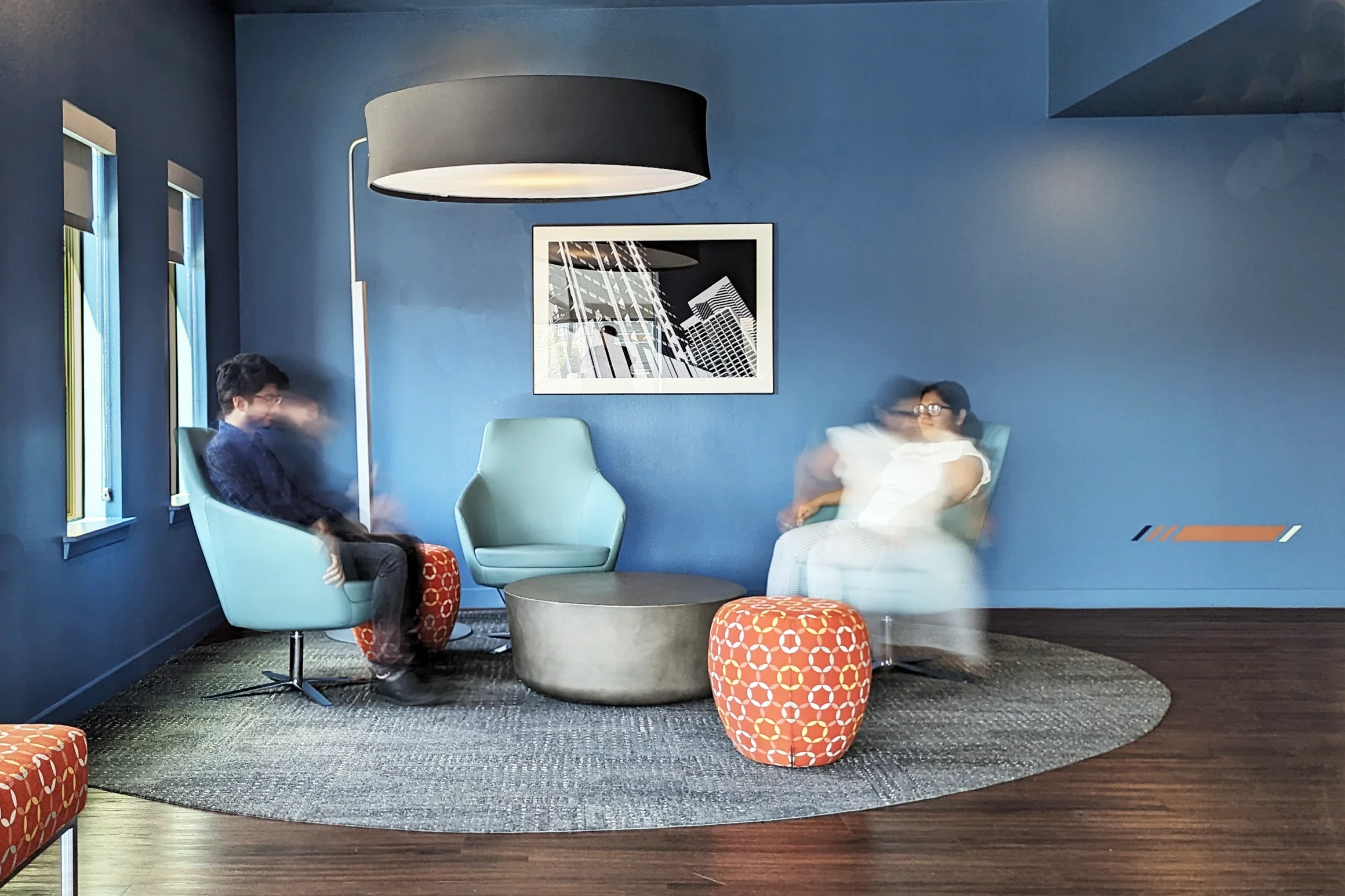 A modern waiting room or lounge with blue walls, a round black coffee table, a large gray drum-shaped ottoman, and two orange patterned ottomans. There are three chairs with two occupied by blurred people, indicating movement. A woman wearing glasses