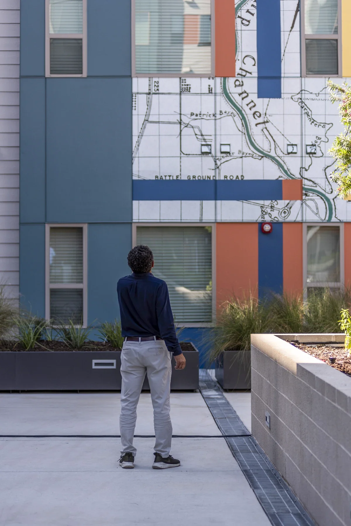 A man standing and looking at a building with a mural that features a map of Channel Lake, with trees and planters nearby.