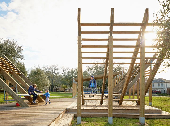 Children playing on a wooden playground structure in a park during the daytime with trees and houses in the background.
