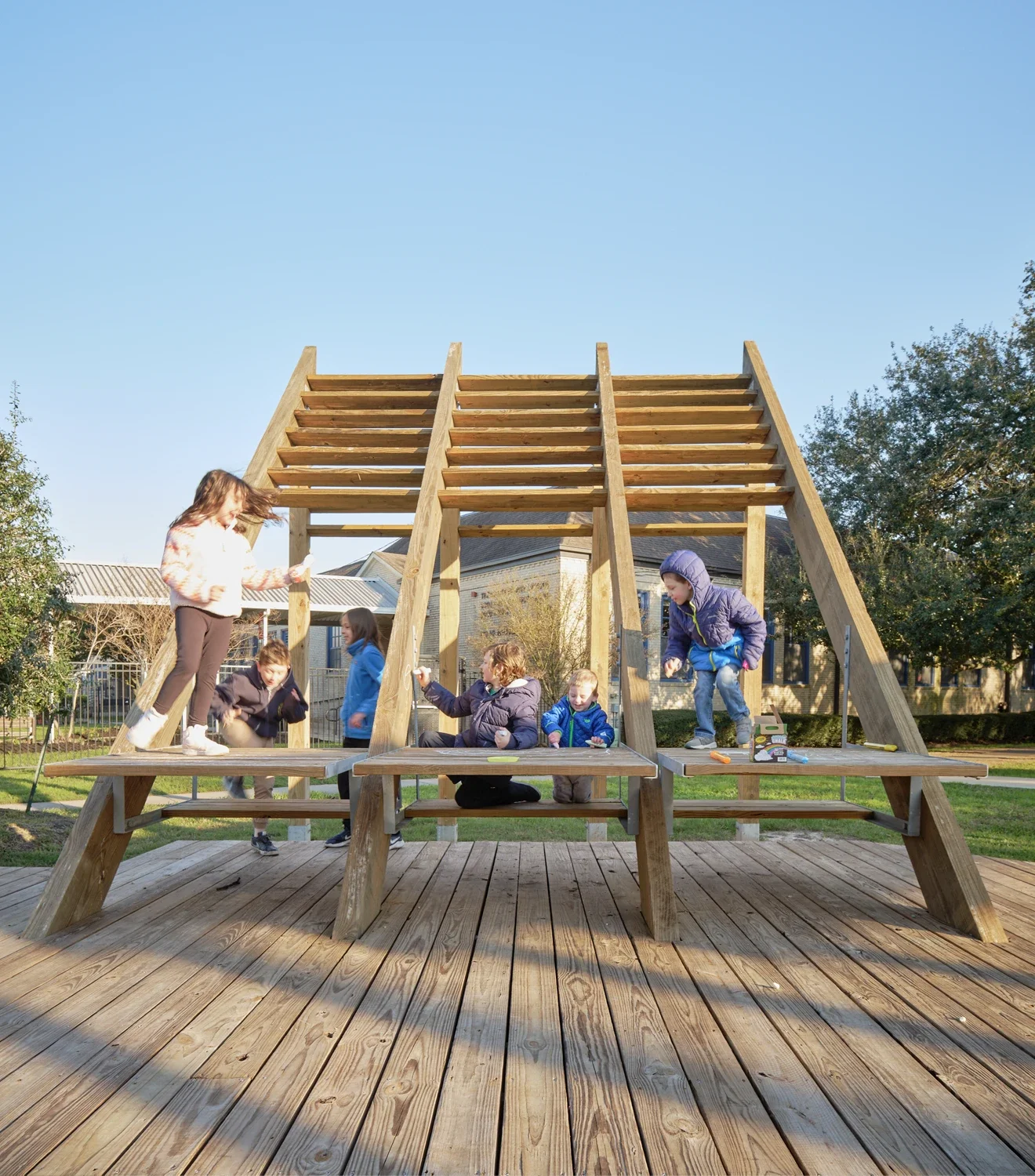 Children playing on a wooden playground structure on a sunny day with a clear blue sky, trees, and houses in the background.