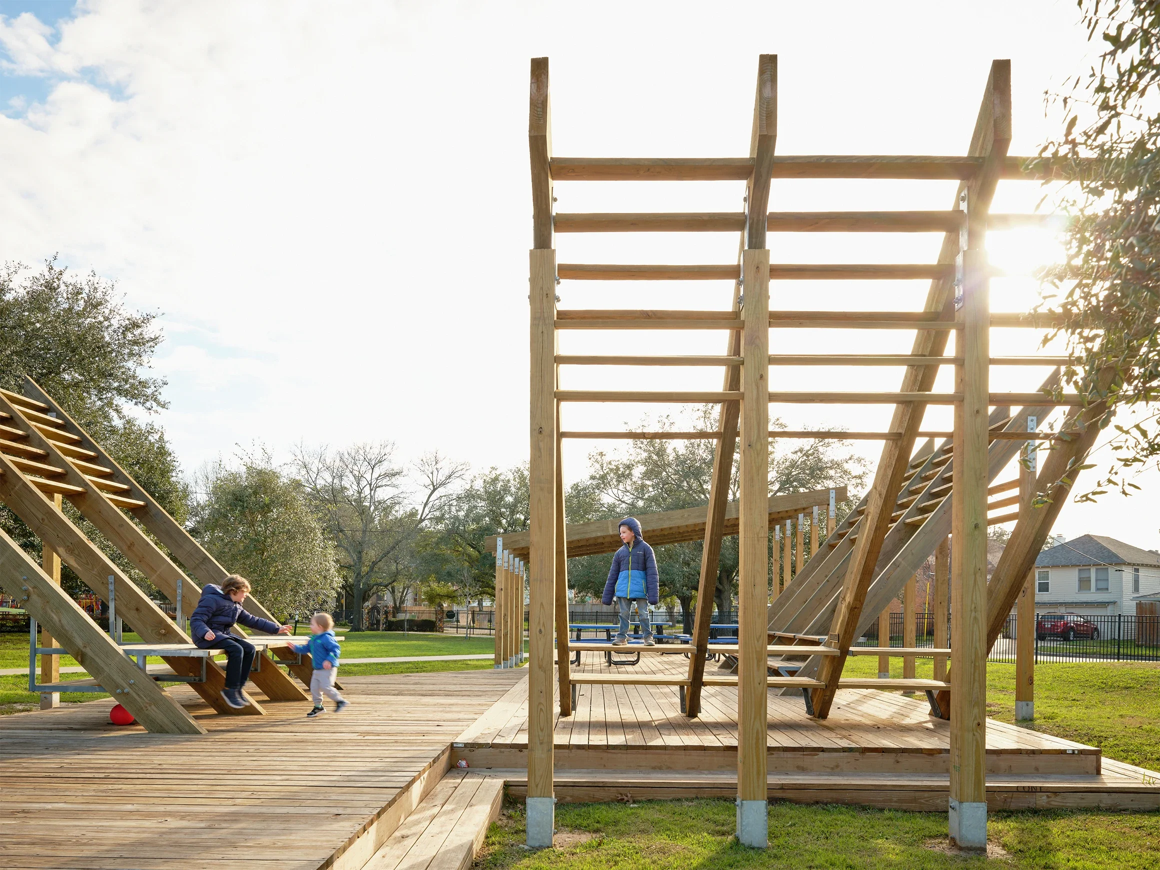 Children playing on a wooden playground structure in a park during daytime, with trees and houses in the background.