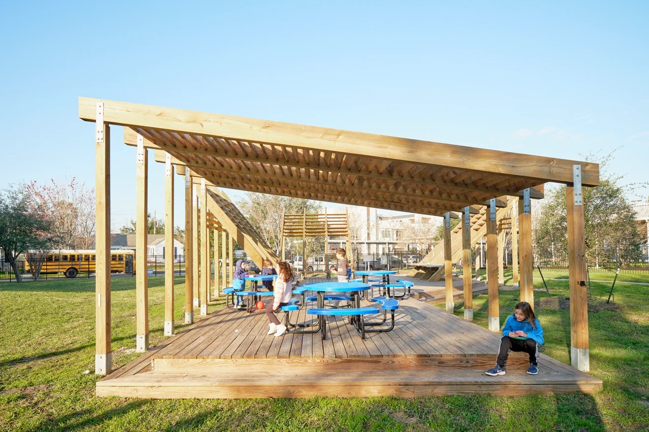 Kids playing on a covered wooden playground with picnic tables in a park.