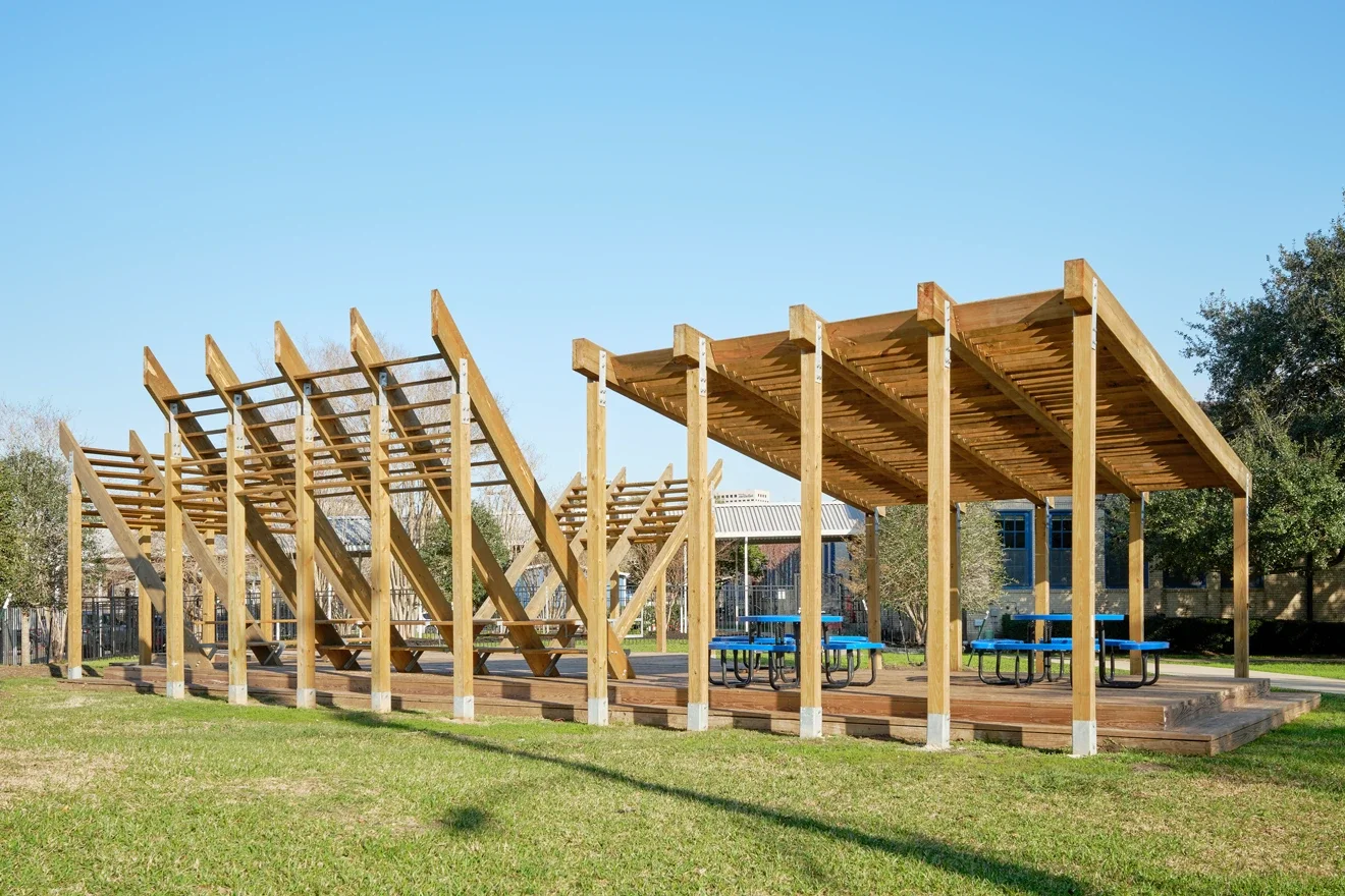 A wooden outdoor pavilion with picnic tables and structures for climbing or playing, situated on a grassy area with trees and buildings in the background under a clear blue sky.