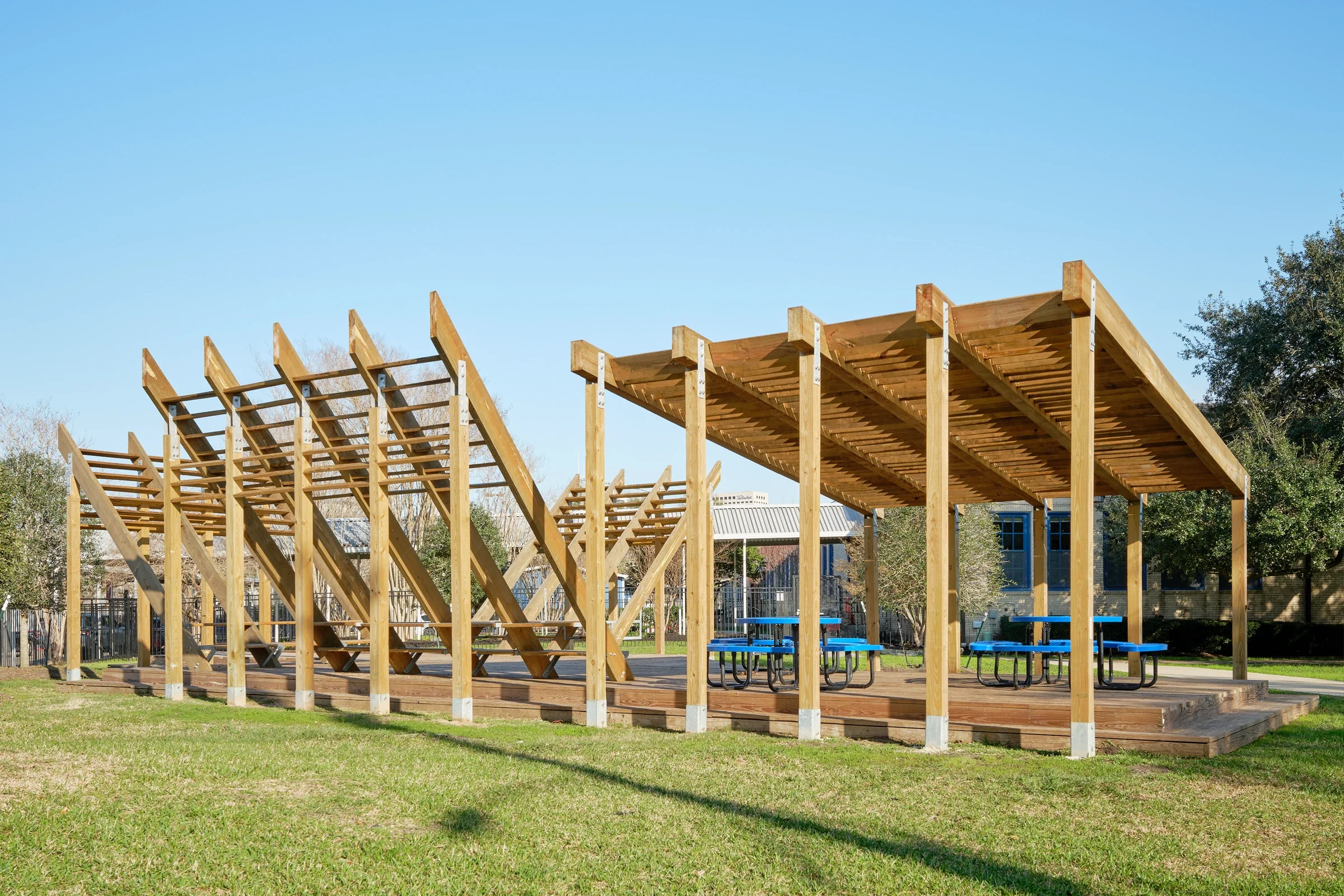 A wooden outdoor pavilion with a slanted roof, set on a grassy lawn under a clear blue sky. The pavilion has picnic tables with blue seats underneath.
