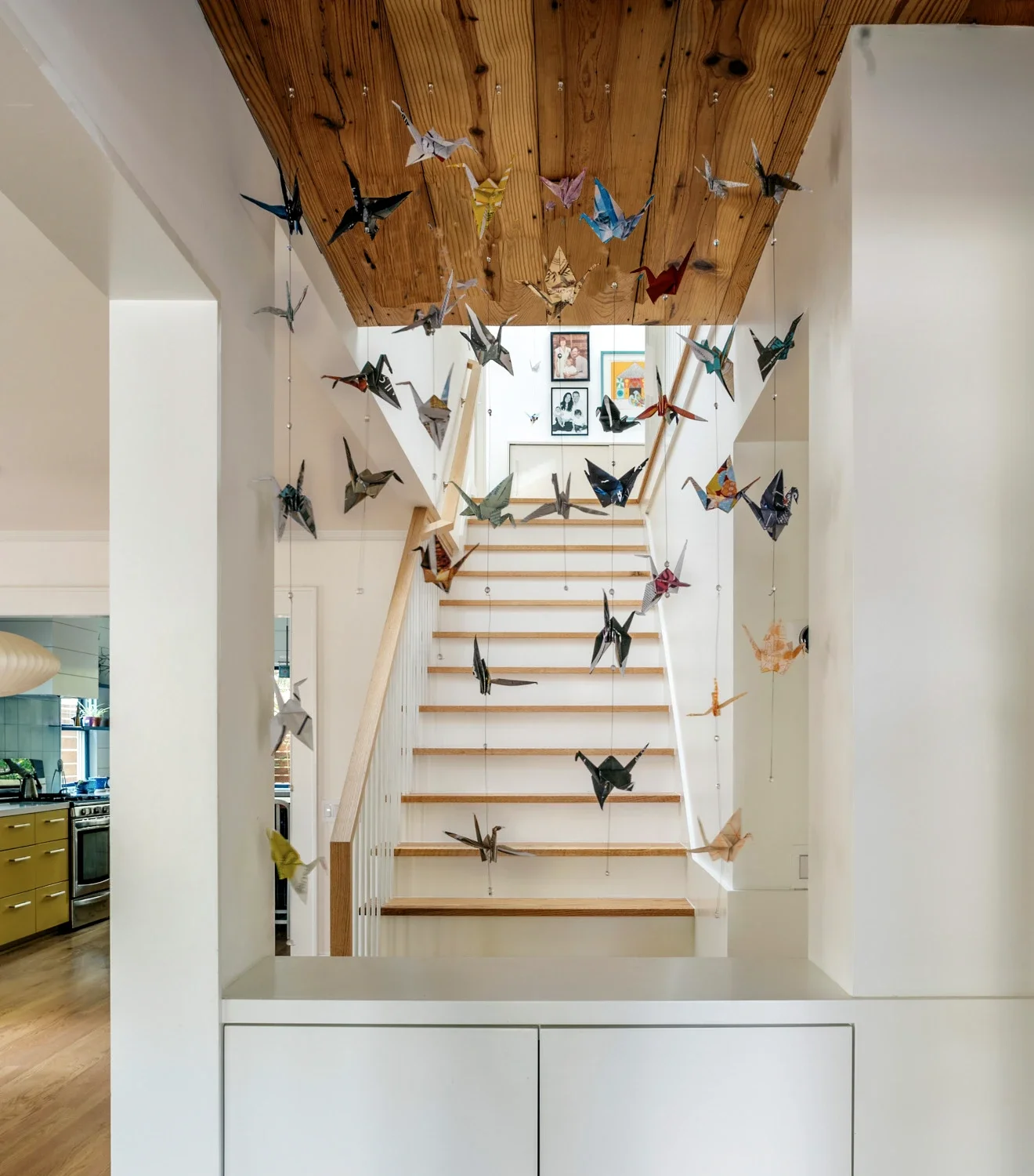 Colorful origami paper cranes hanging from the ceiling above a staircase, with framed photos on the wall at the top of the stairs.