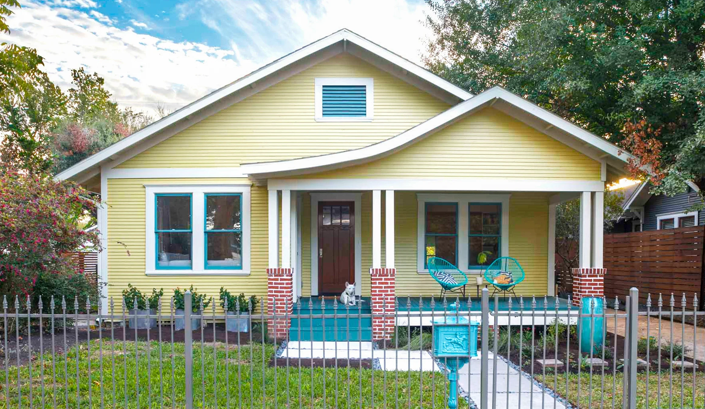 Yellow house with front porch, two turquoise chairs, dog sitting on porch, garden, and fence.