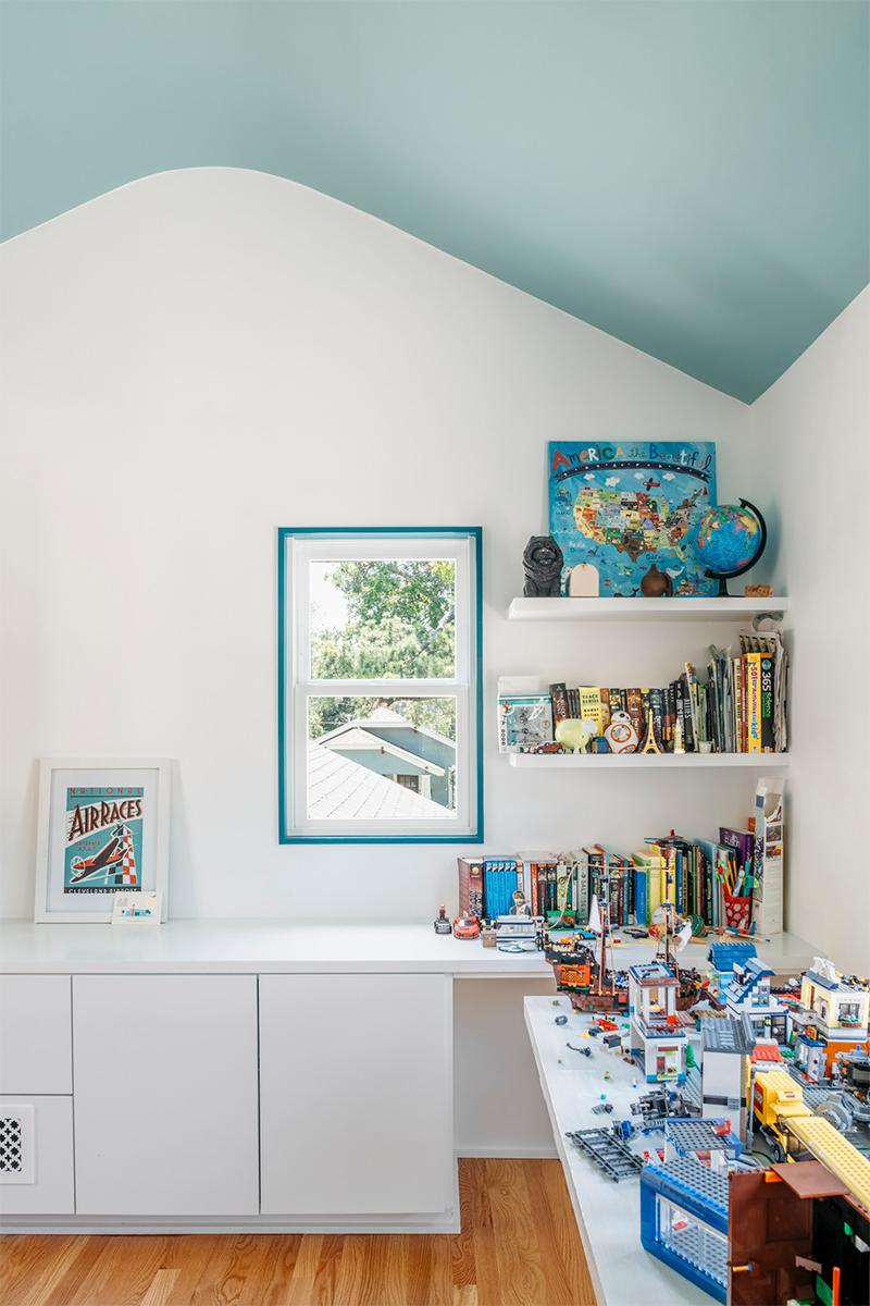 A room with white walls and a sloped ceiling painted light blue. There is a small window showing trees outside. The room has white built-in cabinets and shelves filled with colorful books, toys, and decorations. A LEGO cityscape is built on the white desk or table.
