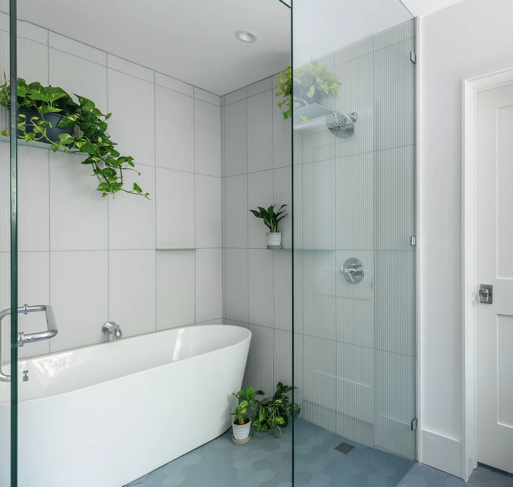 Modern bathroom featuring a white bathtub, glass shower walls, white tiled walls with vertical designs, greenery in potted plants, and a white door.