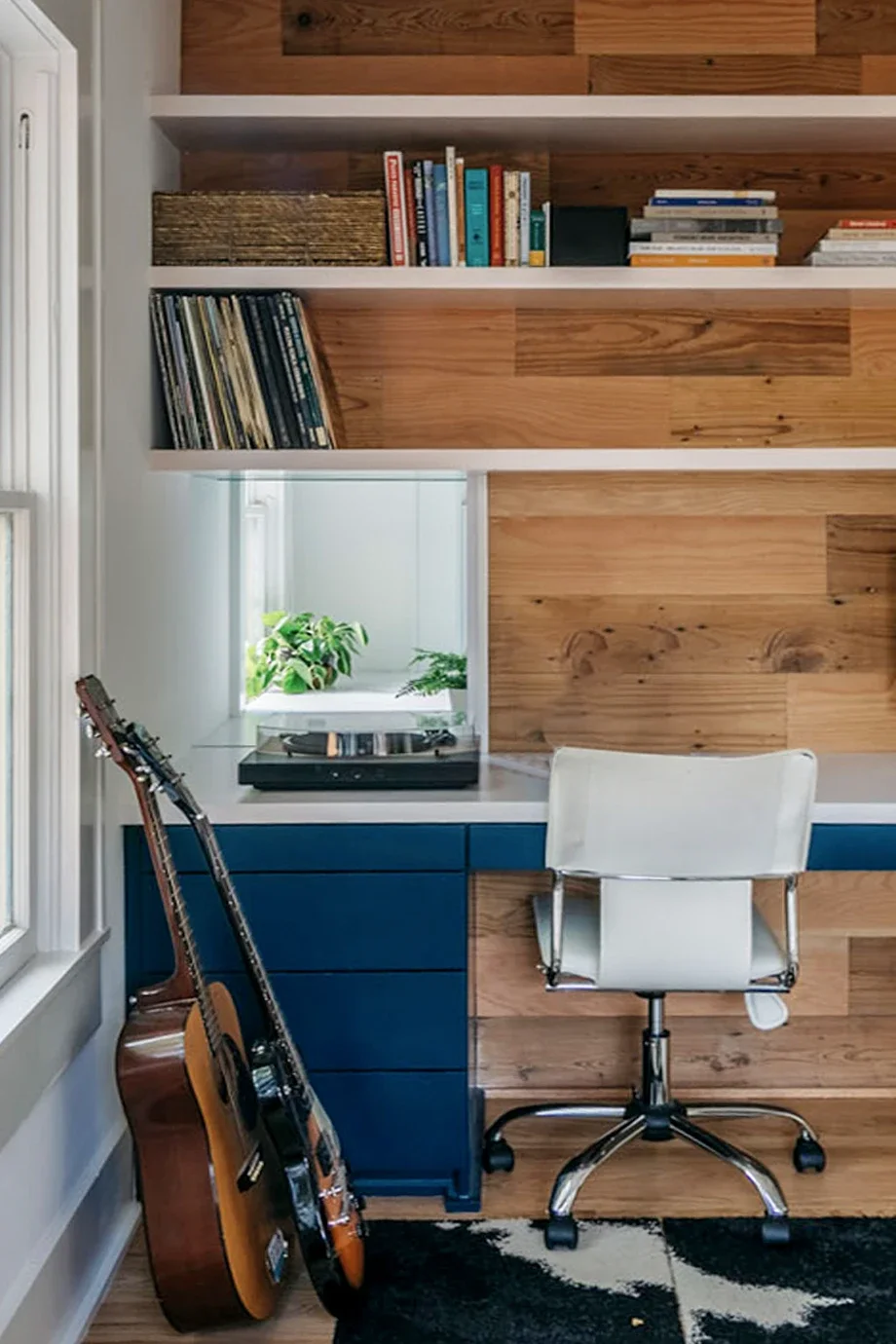 A cozy workspace with a white desk, white swivel chair, blue drawers, and a wooden wall. Guitars lean against the wall, and a window with plants provides natural light.