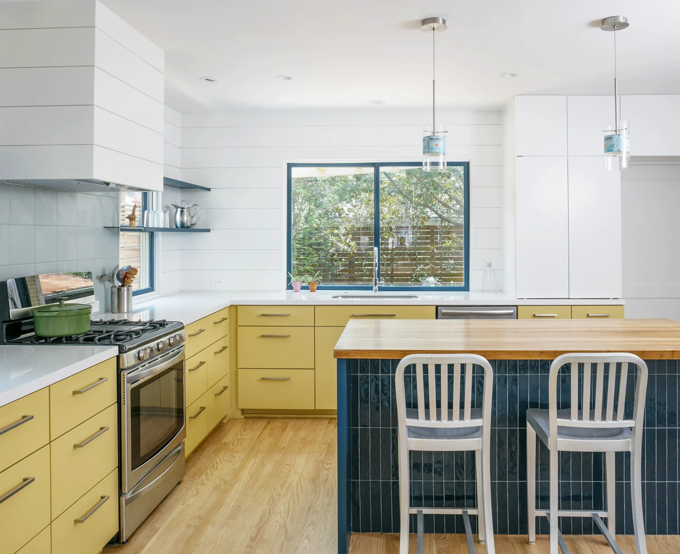 Modern kitchen with yellow cabinets, a white countertop, a stainless steel oven, and a wooden island with blue tile accents, illuminated by natural light from large windows.
