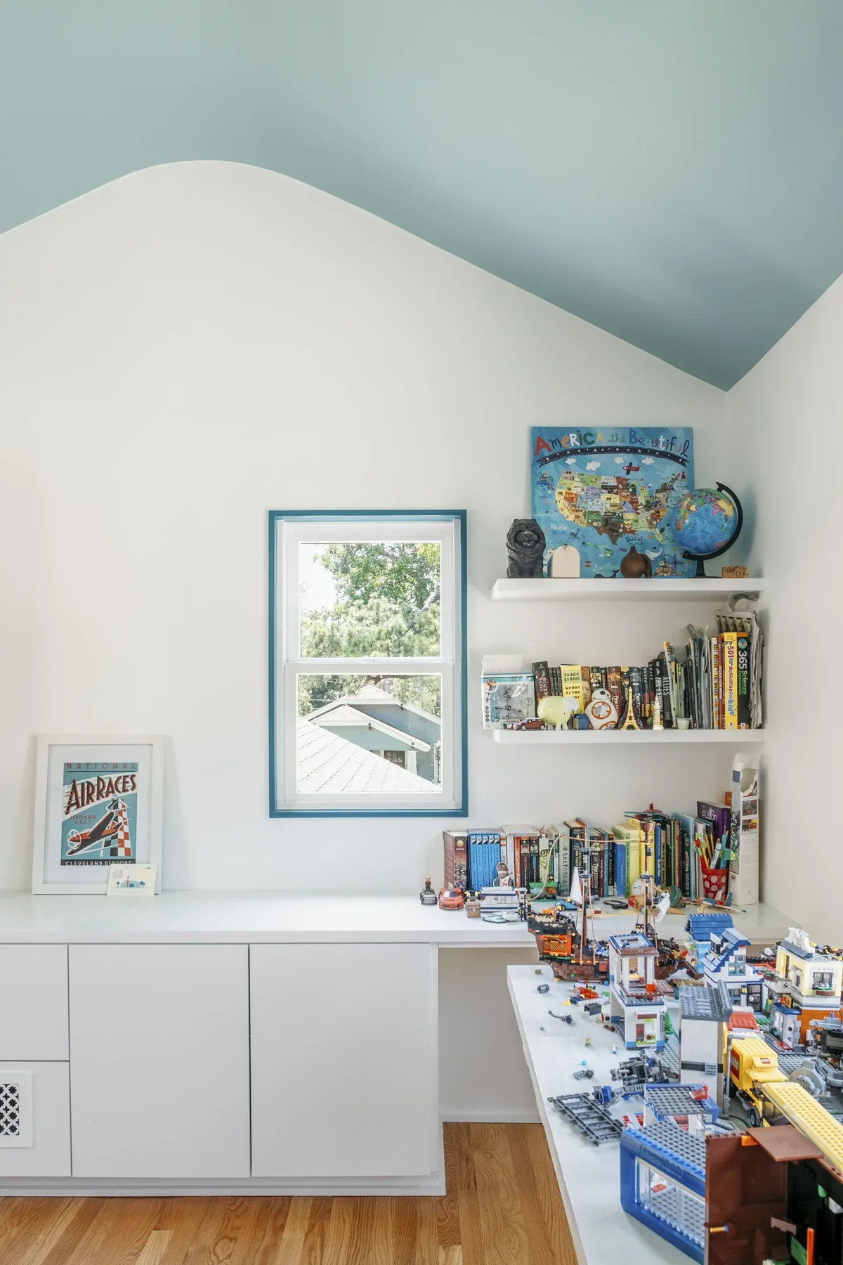 A white built-in bench and bookshelf in a room with a small window and a slanted ceiling painted light blue. The bookshelf contains colorful books, toys, and a LEGO cityscape on a white table in the foreground. A framed poster titled 'National Air Ra