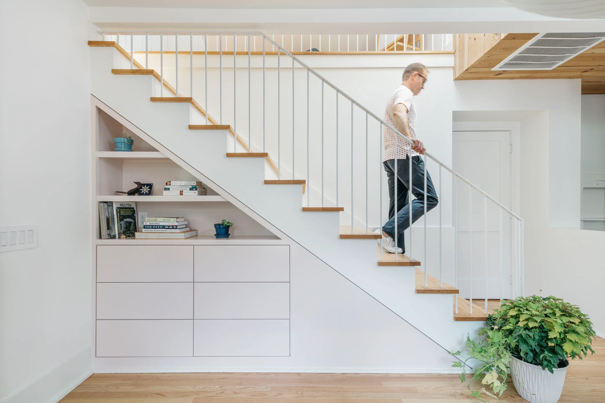 Man walking down a staircase in a modern, minimalist home with white walls, wooden accents, and a large potted plant.