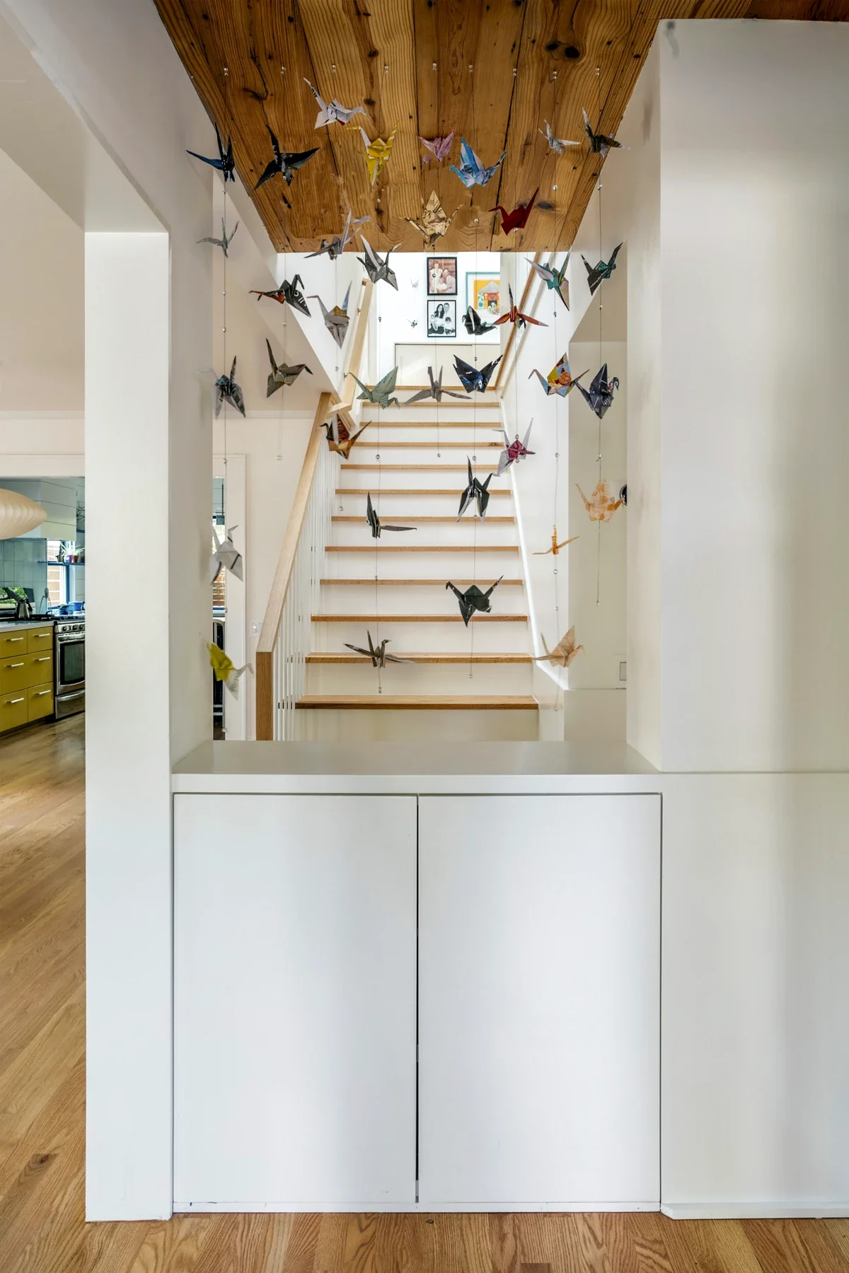 Colorful origami cranes hanging in the doorway of a modern home interior, leading up wooden stairs to a gallery wall of framed pictures.