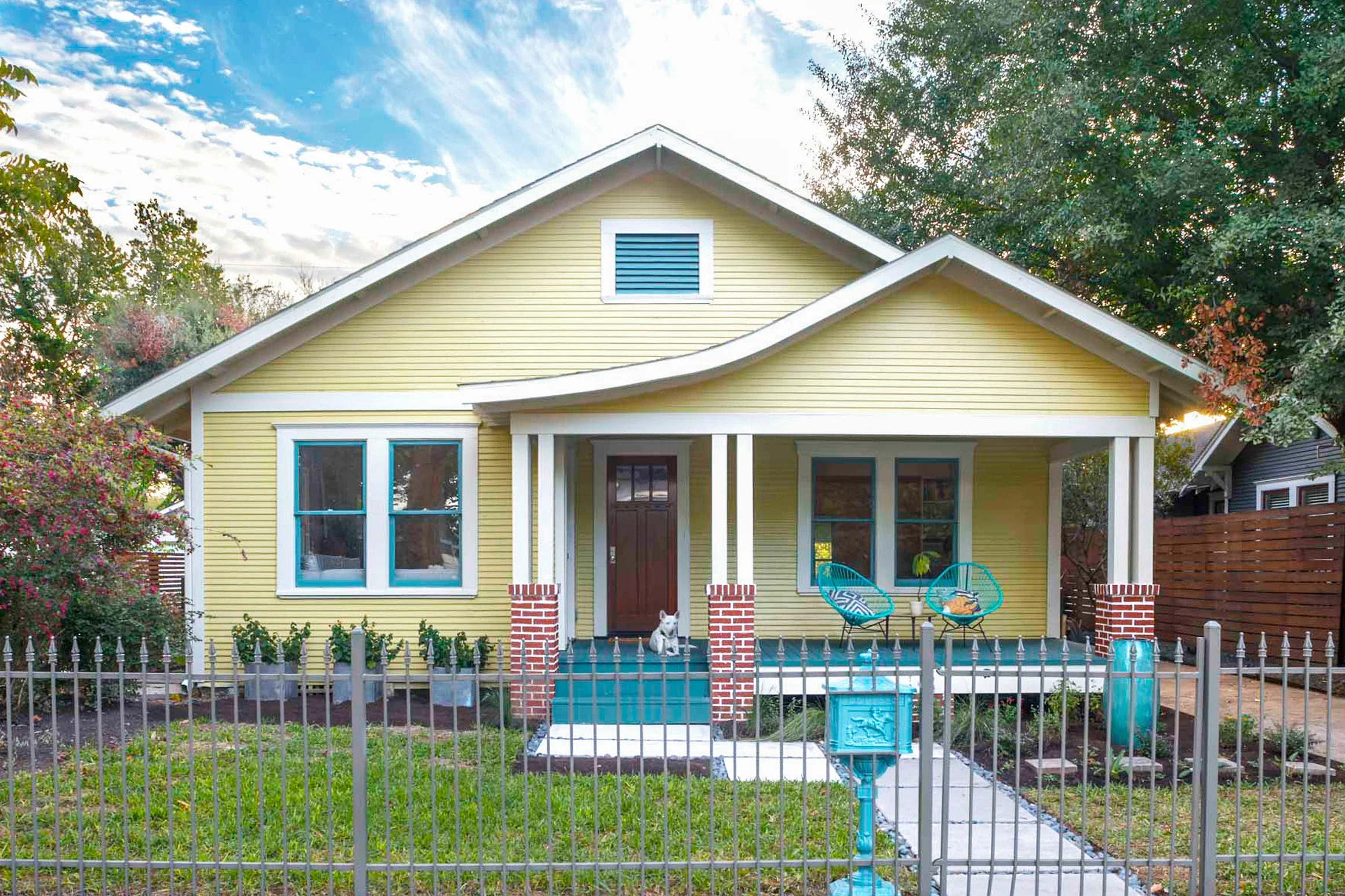 Yellow house with front porch, two chairs, a small dog sitting on the steps, green lawn, trees, and a fence in the foreground, blue sky with clouds in the background.
