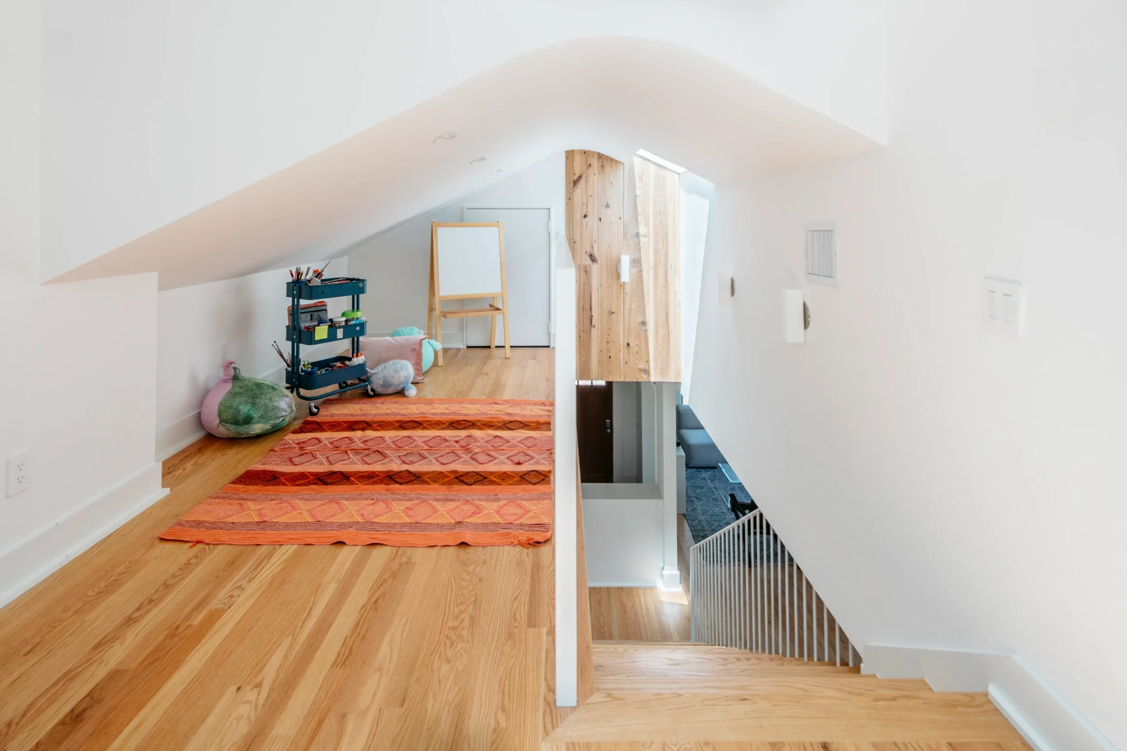 Top view of an upstairs hallway with a staircase leading down to the entryway, featuring white walls, a wooden floor, a colorful orange and red rug, a blue storage cart with art supplies, and a whiteboard on an easel.
