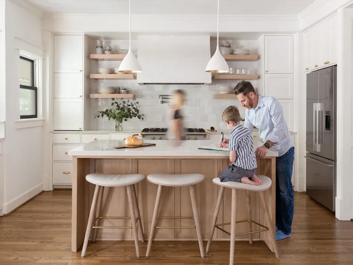A man and a young boy sit on kitchen island stools, with the boy taking notes and the man smiling. In the background, a woman is blurred as she walks through a bright, modern kitchen with white cabinets, open shelves, and a stainless steel refrigerator.