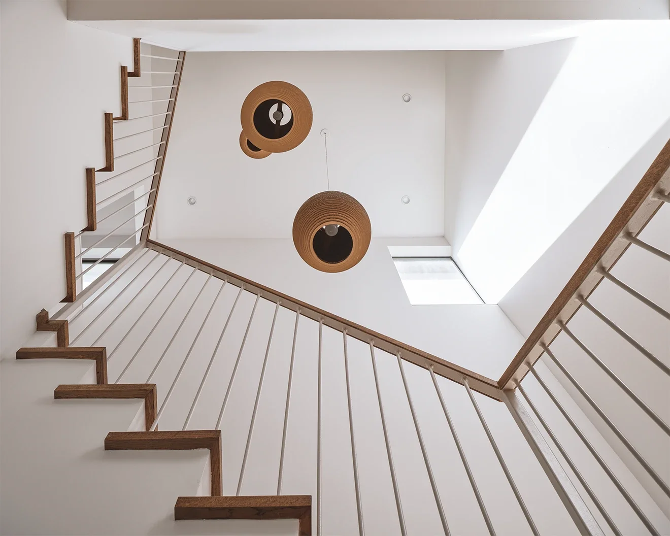 Looking up at a staircase from the bottom, featuring wooden handrails, white balusters, two hanging ceiling lights, and a skylight.