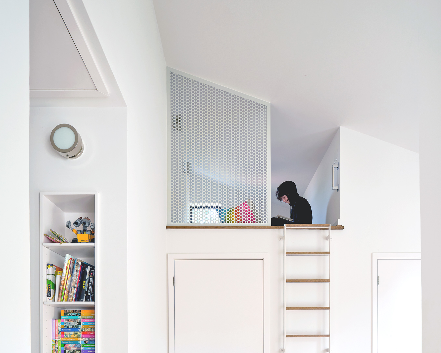 A woman with black hair reading a book on a lofted platform in a white modern room. The room has a small bookshelf filled with books and toys, and a wooden ladder leading up to the platform. There's a circular light fixture on the wall and a decorative white perforated panel.