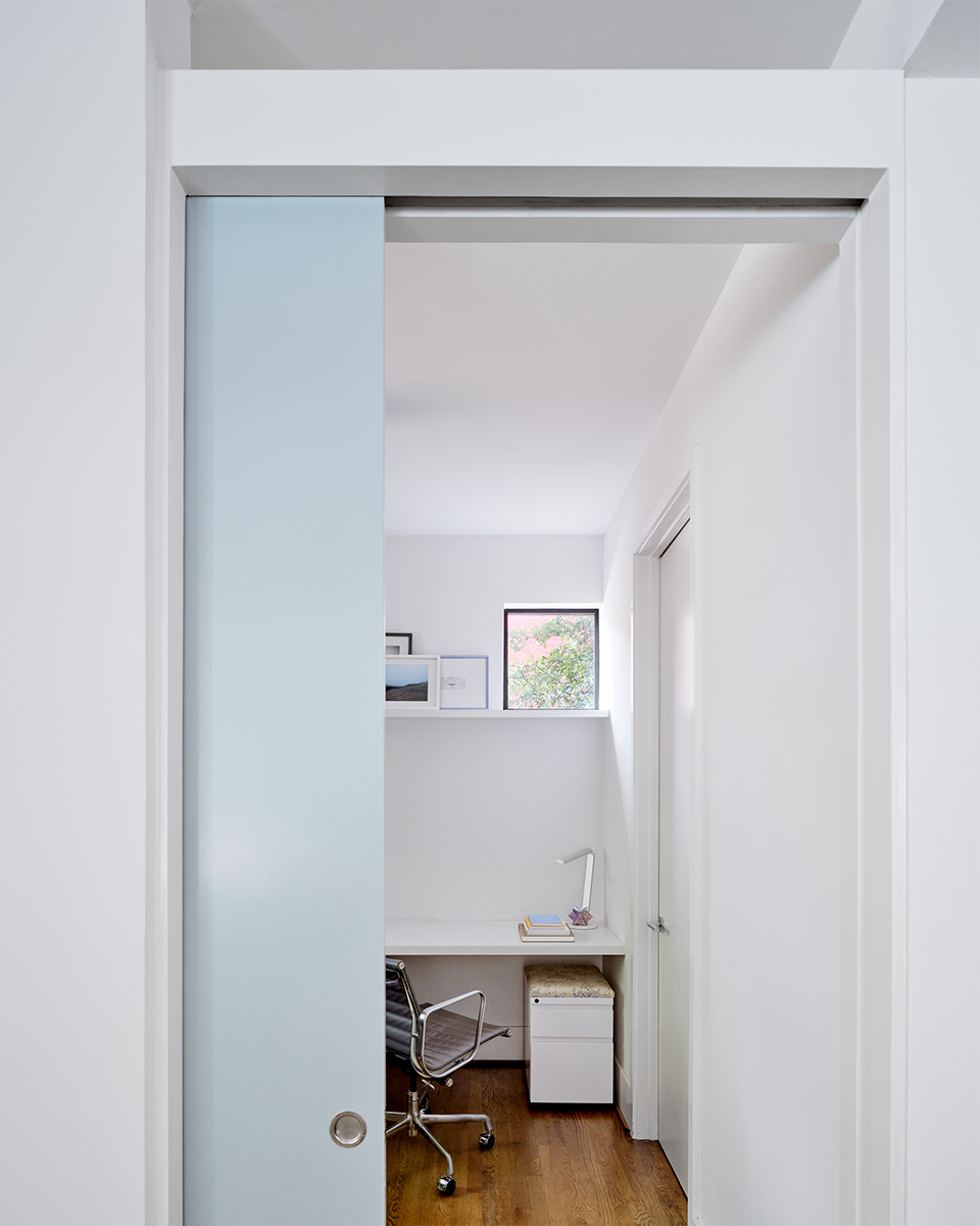 Small home office with a sliding frosted glass door, a white built-in desk, a swivel chair, a window with framed pictures on the ledge, and a small storage cabinet under the desk.