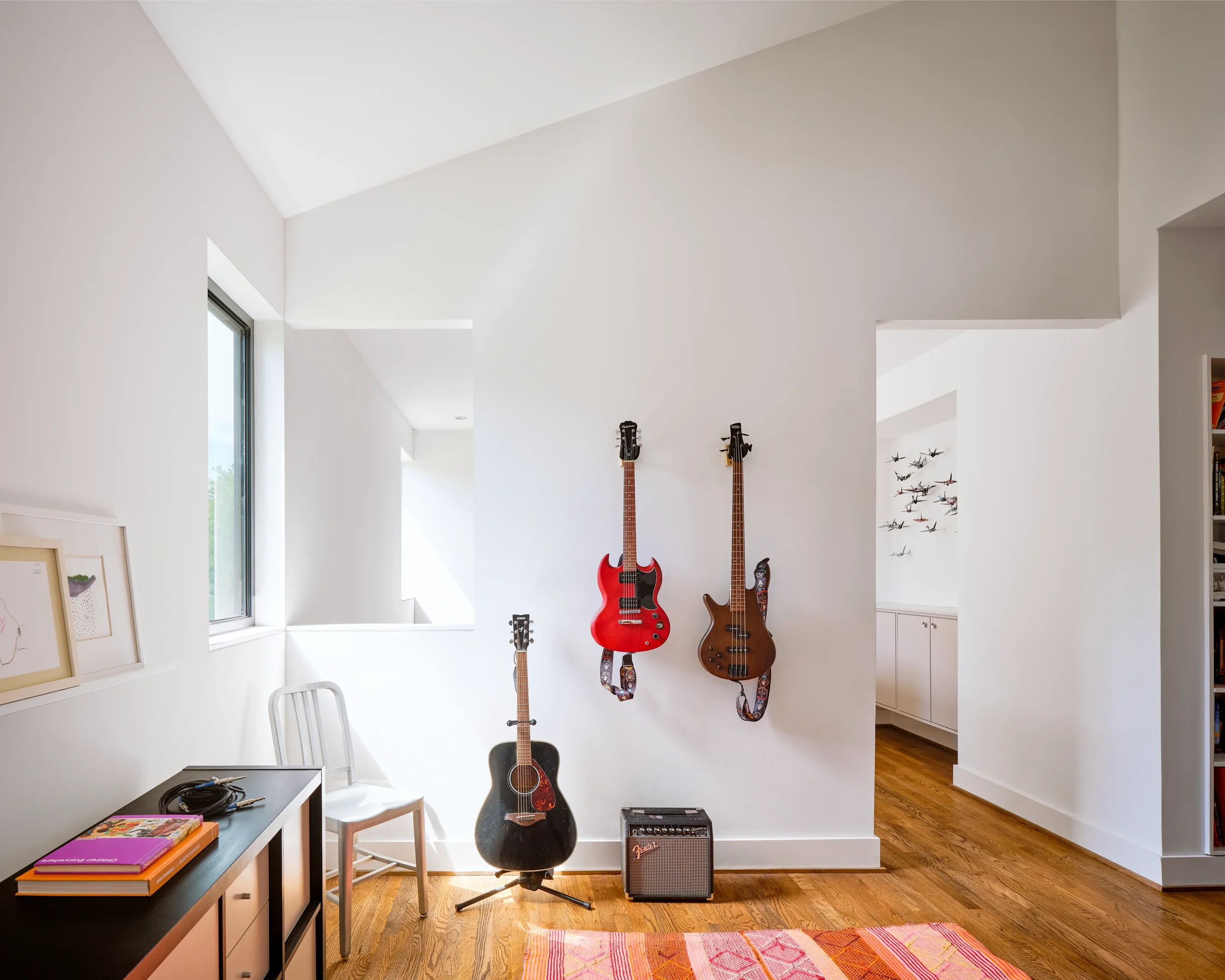A room with white walls, wooden floor, three guitars hanging on the wall, a black amplifier, a black guitar on a stand, a white chair, and a side table with books and headphones. Sunlight coming through a window on the left.