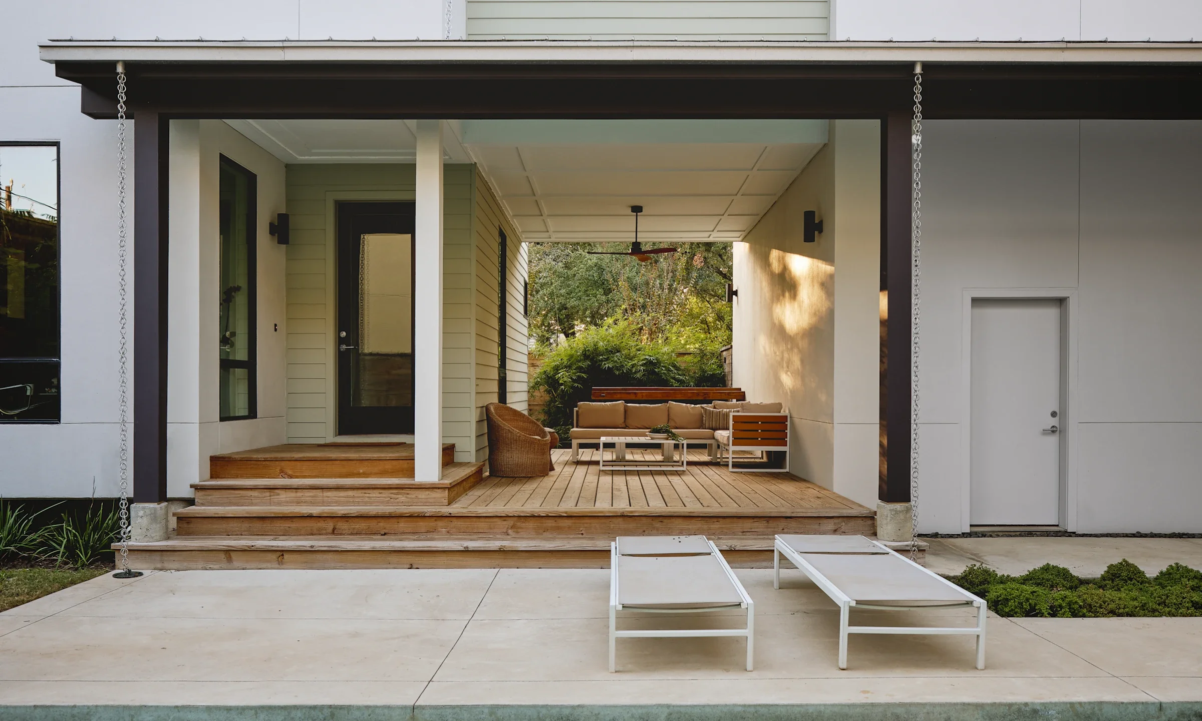 Modern house patio with wooden deck, outdoor seating with a sofa and chairs, two white lounge chairs in front, surrounded by greenery, with a black ceiling fan and a ceiling light.