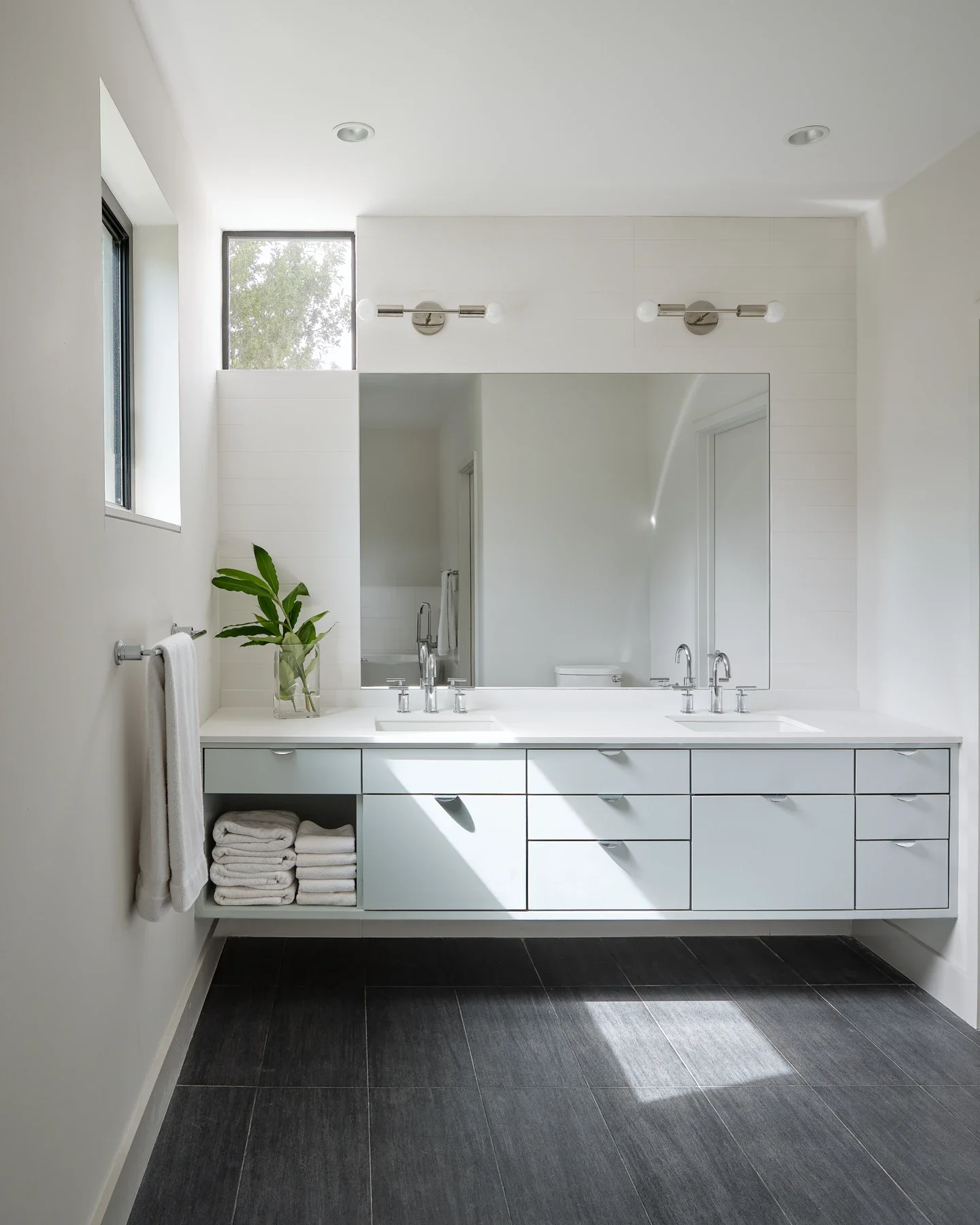 Modern white bathroom with double sink vanity, large mirror, window, black tiled floor, and natural light.