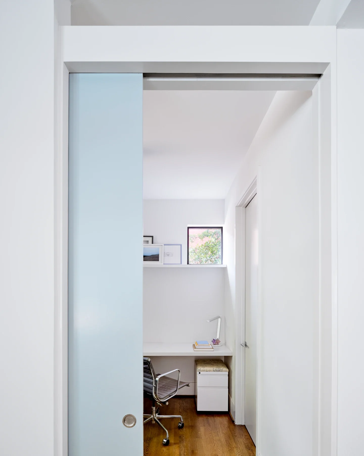 Small minimalist home office with white walls, a white desk, books, a modern desk lamp, and an office chair, with a window showing greenery outside.