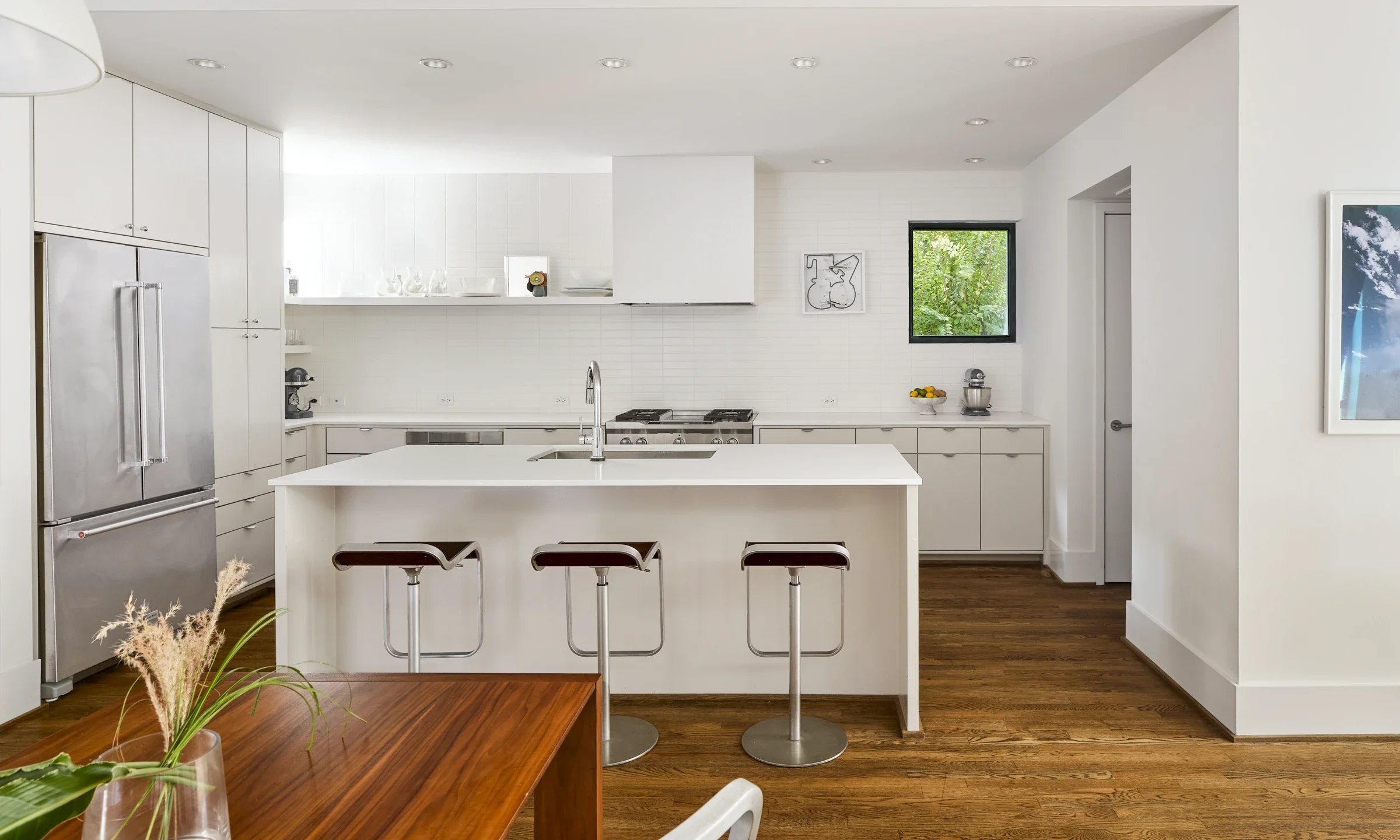 Modern white kitchen with wooden flooring, center island with three brown bar stools, stainless steel refrigerator, and small window showing greenery outside.